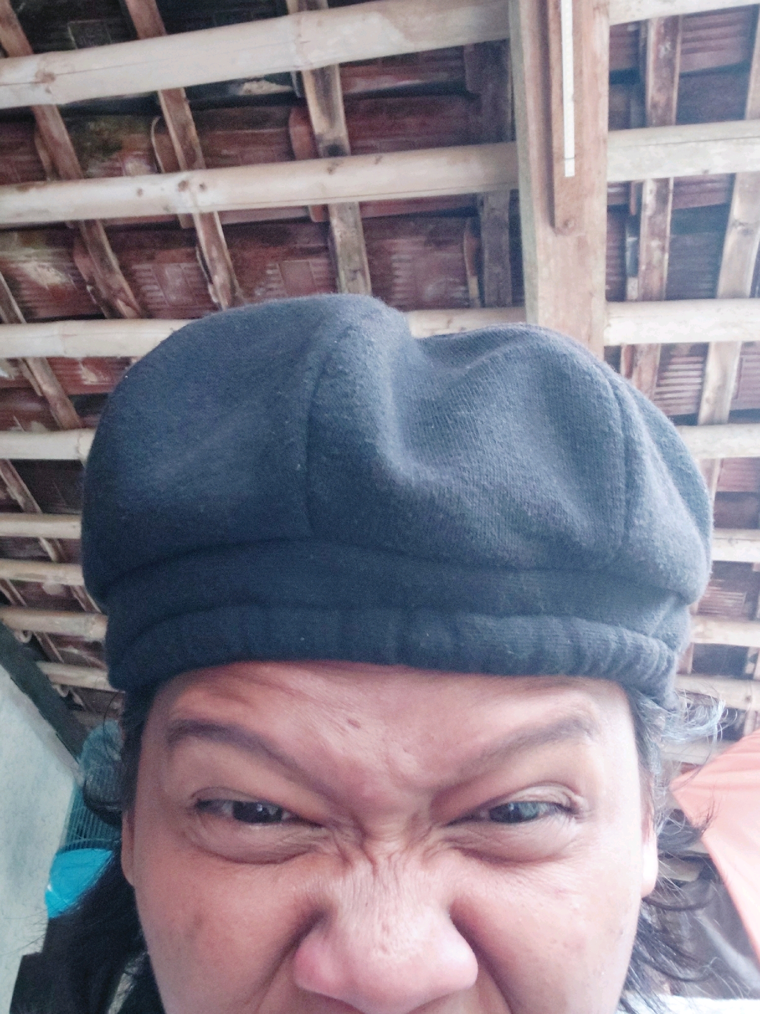 The image shows a close-up of a person's face, wearing a dark beret. The person's eyes are scrunched up, giving them a grimacing expression. The background consists of wooden beams and planks, suggesting an indoor or covered outdoor space with a rustic roof structure. There is no discernible urban environment or reference to Bangkok, Indonesia.