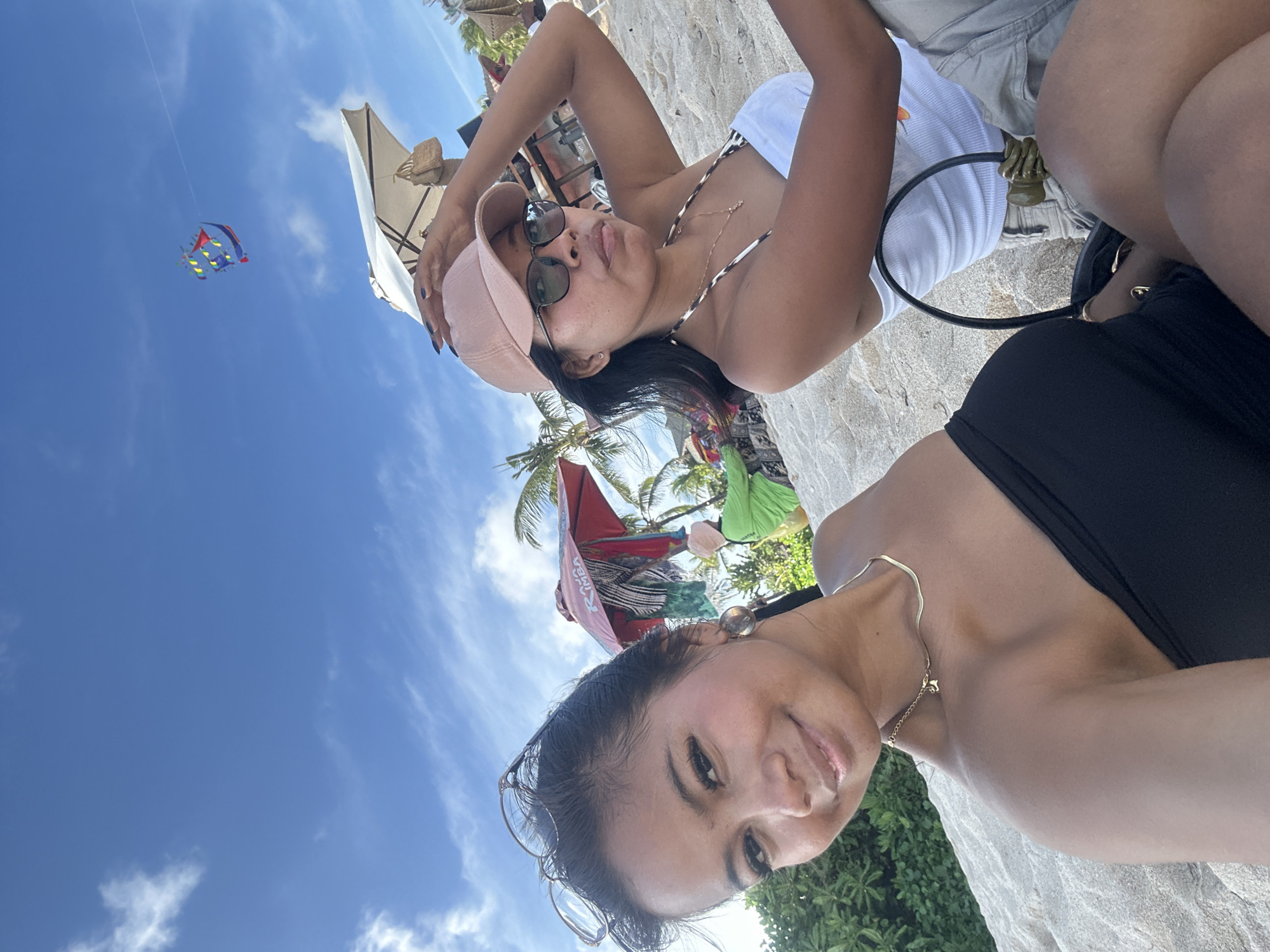 This outdoor photo, taken in Kuta Selatan, Indonesia, features two women relaxing on what appears to be a sandy beach or resort area under a clear blue sky. The image is oriented with the sky on the left.
In the foreground, a woman with dark hair, wearing a black top and a thin gold-colored necklace with a small charm, smiles gently towards the camera. She has glasses resting on her head. Behind her, a second woman with dark hair is visible, wearing a light pink baseball cap, sunglasses, and a white top with a patterned (possibly leopard print) strap. She is posing casually, looking up and slightly away from the camera, with a playful expression. Parts of another person's legs, clad in shorts, are visible between the two women, suggesting a third individual or the posture of the second woman.
The background reveals a vibrant blue sky with scattered white clouds, where a colorful kite (featuring red, blue, yellow, and green sections) flies high. Several beach umbrellas or tents, including a light-colored one and a red one with some indistinct text, are visible among palm trees, indicative of a tropical setting. The ground beneath the subjects is sandy.
The scene depicts a sunny day, likely midday or early afternoon, characterized by a relaxed and leisurely atmosphere. The women appear to be enjoying their time outdoors, possibly at a beach or resort in Indonesia.