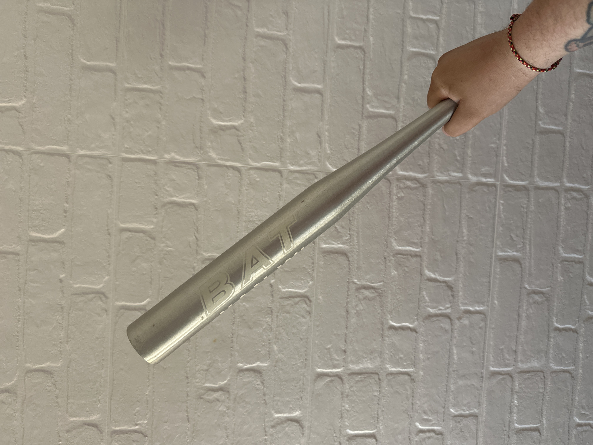 A person is holding a silver-colored metal baseball bat in their right hand. The bat is positioned diagonally across the frame, with the grip end near the person's hand and the barrel end pointing towards the lower left corner. The word "BAT" is etched on the side of the bat. The person is wearing a beaded bracelet on their right wrist. The background is a white textured brick wall. There is no indication of this image being in Sant Feliu de Guíxols, Spain.
