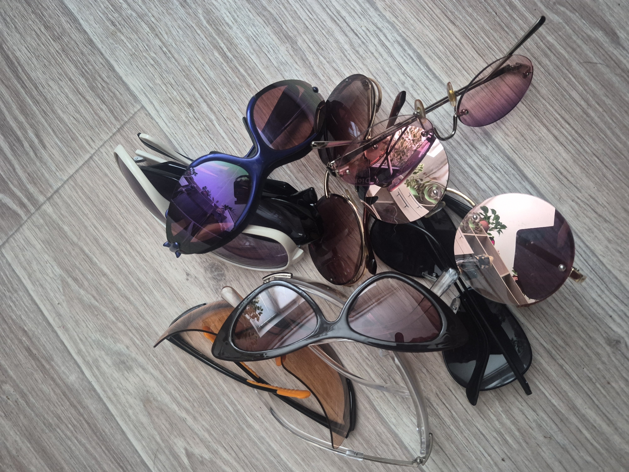 A pile of various sunglasses is scattered on a wooden floor. The sunglasses have different frame colors and lens tints, including blue, black, and rose gold. Some lenses are reflective and show reflections of what appears to be an indoor environment with plants and furniture. The wooden floor has a light gray, distressed finish with visible wood grain. The lighting suggests it is daytime, and the scene appears to be indoors, possibly a home or a store. There is no apparent activity taking place other than the arrangement of the objects. No people are visible. No text is clearly legible on the sunglasses.