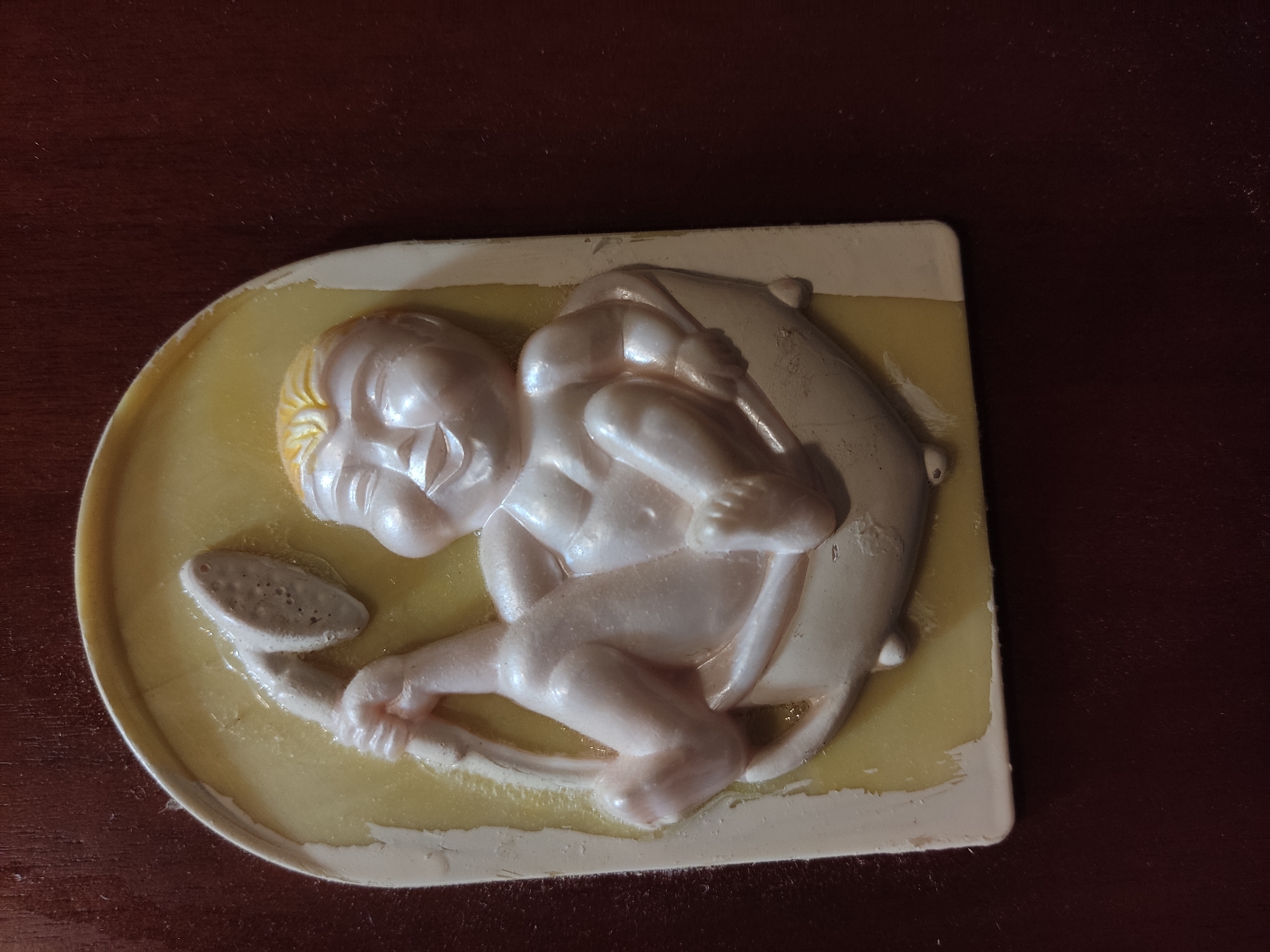 A white, pearlescent bas-relief plaque depicting a cherubic baby sitting in a bathtub holding a shower head is affixed to a dark wood surface. The plaque has a pale yellow background and an irregular white border. The baby is depicted with chubby cheeks, closed eyes, and a wide smile, with its legs bent and arms outstretched. The shower head is held in the baby's right hand, and the left hand rests on its thigh. The plaque is slightly chipped and worn, particularly along the edges. The dark wood surface appears to be a wall or furniture, with a subtle grain pattern. The image is taken from a slightly overhead angle, with the plaque positioned in the lower left quadrant of the frame. The scene appears to be indoors. There is no discernible information to determine the location in the city of Noginsk, Russia.