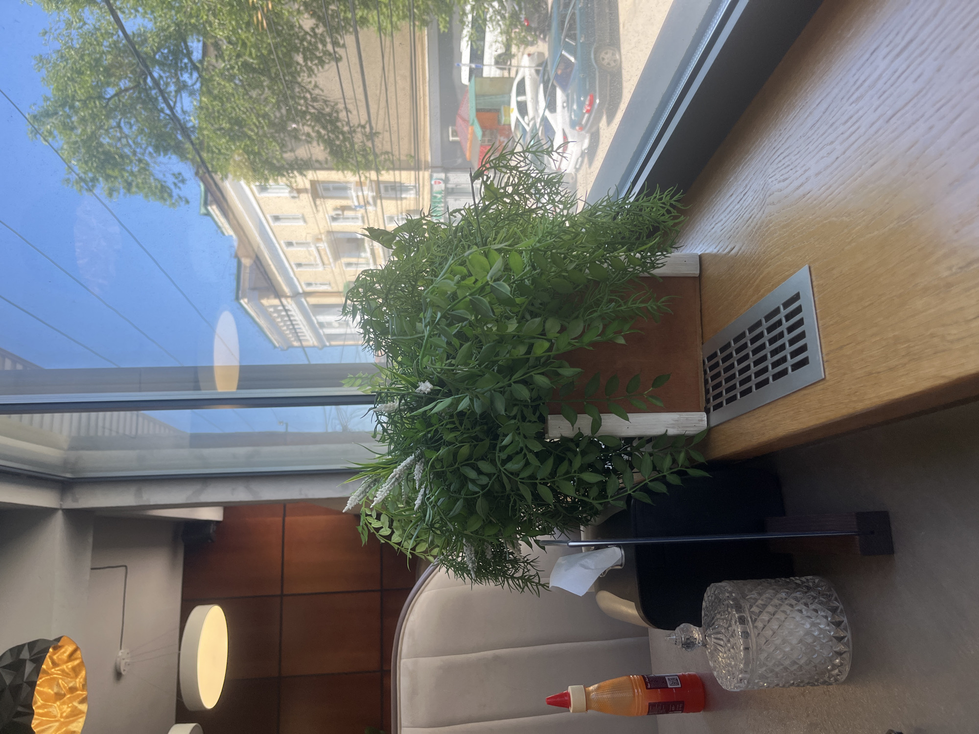 The image, captured in Kaspiysk, Russia, presents a brightly lit interior scene looking out onto an urban environment through a large window. In the foreground, a vibrant artificial plant with numerous green leaves and some white floral accents is prominently displayed in a rectangular wooden planter on a light-colored wooden windowsill or counter. To the right of the plant, a silver-colored vent is integrated into the wooden surface.
Through the window, a clear blue sky and abundant green trees are visible, suggesting a sunny day. A light-colored, multi-story building with numerous windows stands across the street, below which several cars are parked. Overhead power lines or utility wires are also visible against the sky. Reflections on the glass add a shimmering quality to the outdoor view.
The interior space includes a segment of a comfortable, upholstered beige seating, likely part of a booth or armchair. On the surface next to the seating, a decorative clear glass jar with a patterned surface and a lid is present. An orange-red squeeze bottle with a red cap, possibly containing sauce or condiment, also sits on the surface, displaying a QR code and unreadable text on its label. A white tissue is visible, suggesting a tissue holder nearby. The background reveals modern interior design elements, including varied wall textures, a circular light fixture, and a geometric gold and black light. The overall scene suggests a cafe, restaurant, or lounge setting during daytime.