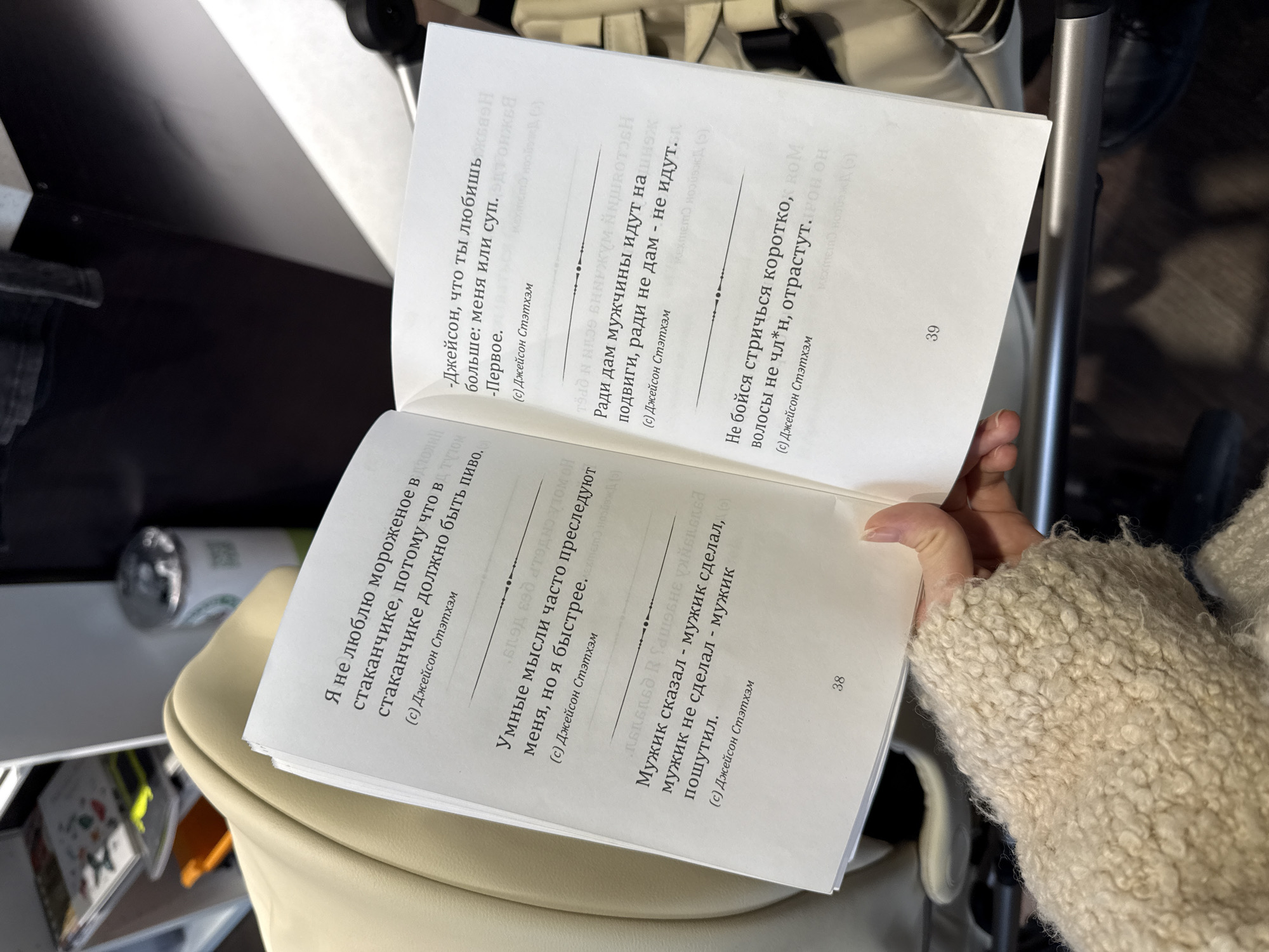 A close-up, slightly tilted shot shows a person's hand holding an open book. The book is white with black text, featuring quotes in Russian, all attributed to "Джейсон Стэтхэм" (Jason Statham).
The left page (page 38) displays three quotes:
1. "Я не люблю мороженое в стаканчике, потому что в стаканчике должно быть пиво." (Translation: "I don't like ice cream in a cup, because there should be beer in a cup.")
2. "Умные мысли часто преследуют меня, но я быстрее." (Translation: "Smart thoughts often chase me, but I am faster.")
3. "Мужик сказал - мужик сделал, мужик не сделал - мужик пошутил." (Translation: "A man said - a man did, a man didn't do - a man joked.")
The right page (page 39) also shows three quotes:
1. "-Джейсон, что ты любишь больше: меня или суп. -Первое." (Translation: "-Jason, what do you love more: me or soup. -The first.")
2. "Ради дам мужчины идут на подвиги, ради не дам - не идут." (Translation: "For ladies, men go on feats; for those who won't give, they don't go.")
3. "Не бойся стричься коротко, волосы не чл*н, отрастут." (Translation: "Don't be afraid to cut your hair short, hair isn't a dick, it will grow back.")
The person's hand, partially visible on the bottom right, appears to be holding the right page. The arm is covered by a light-colored, fluffy, possibly wool or teddy-fabric sleeve. In the background, out of focus, are parts of what appears to be a light-colored stroller or car seat, and some indistinct items, possibly shelves or other furniture. The lighting suggests an indoor setting. The book pages are numbered 38 and 39 at the bottom of each page.