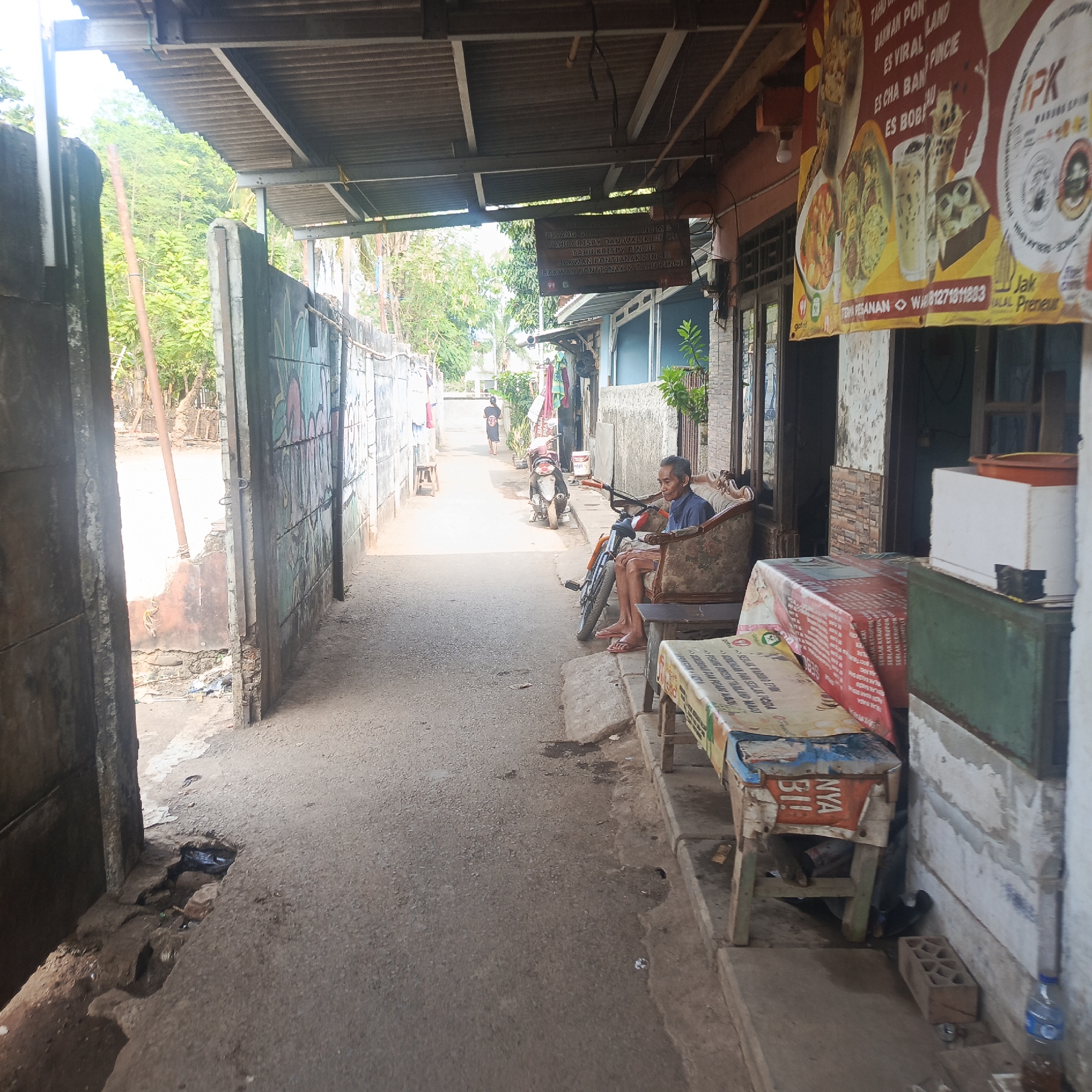 The image depicts a narrow, paved alleyway in a South Jakarta, Indonesia neighborhood. A corrugated metal roof supported by metal beams extends over the alley, providing shade. On the right side of the alley, a building with a slightly aged facade has a prominent banner displaying food and drink items in Indonesian, including "PISANG GORENG KETAN HITAM," "TAHU CRISPY," and "ES BOBA." Next to the building, a man sits on an ornate, upholstered armchair placed on a concrete platform. A motorcycle is parked in front of the armchair. A wooden bench covered with a patterned cloth, also featuring text, is situated in front of the man. Further down the alley, a person is walking away from the camera. On the left side of the alley, a weathered concrete wall with graffiti is visible, leading to an opening that reveals trees and vegetation. The ground in the alley is worn and shows signs of age, with some debris and a small puddle of water in a cracked section of the pavement near the left wall. The scene suggests a casual, local commercial or residential area.