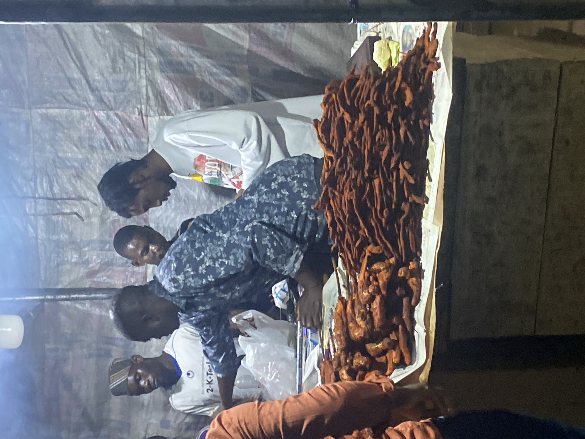 In the city of Karum, Nigeria, at a nighttime market stall, four men are gathered. The primary focus is on a man in the foreground, wearing a dark, patterned shirt, who is actively handling a pile of fried food, possibly plantains or yams, presented on a table covered with paper. To his left, another man, partially visible and wearing an orange shirt, stands. Behind the man in the patterned shirt, two other men are visible: one in a white t-shirt with a logo and graphic print, and another to his left wearing a white t-shirt with the text "2-K-Tech" and a blue rimmed hat. The background consists of a textured, possibly tarp-like material, illuminated by an overhead light source, suggesting an outdoor or covered market setting. The overall scene depicts a food vendor interacting with his products and potentially customers in a bustling nighttime environment.
