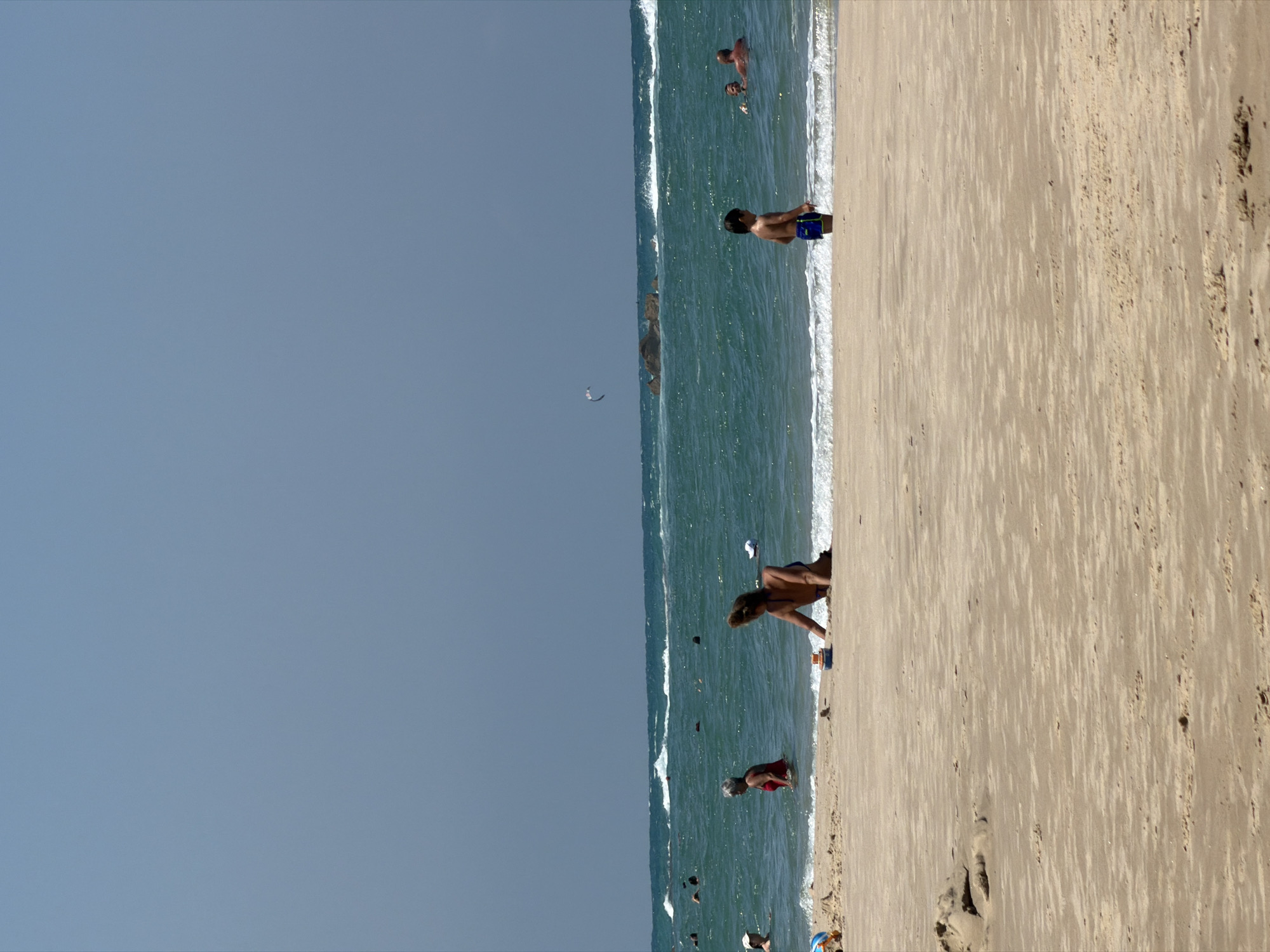 A wide, sandy beach in Bat Yam, Israel, is occupied by a few people on a sunny day with a clear, pale blue sky. In the foreground, the sand is rippled by wind and waves. A woman is partially buried in the sand in the middle ground, leaning back with her head and arms visible. A small, colorful toy boat sits near her. Further out in the turquoise ocean, a young boy in blue swim trunks stands near the shore. Several other people are scattered in the water, swimming or wading. In the distance, a kite flies in the sky above dark rock formations in the choppy waves.