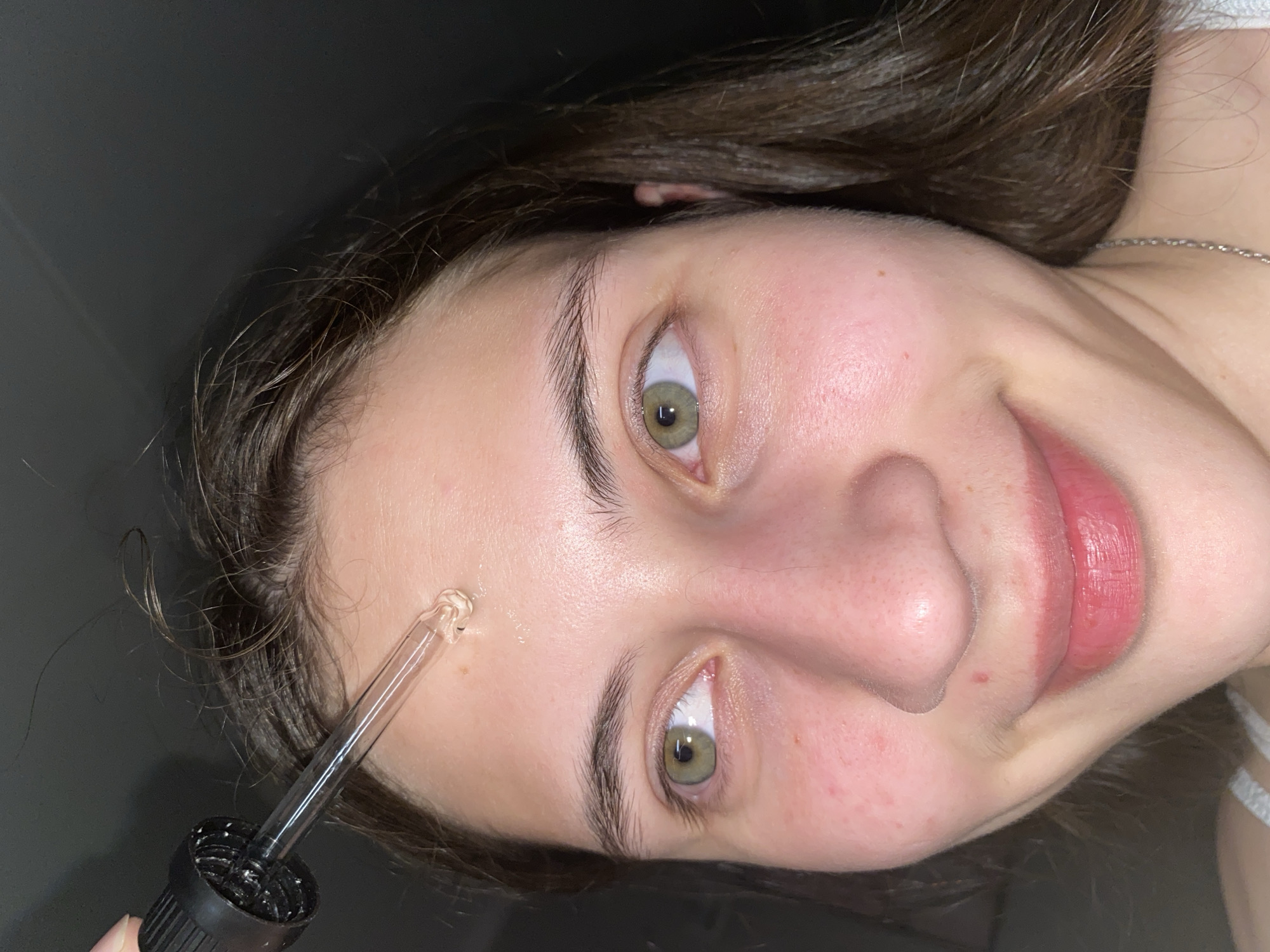 The image shows a close-up view of a young woman's face as she applies a clear liquid serum from a dropper onto her forehead. The woman has light skin with some redness, light brown hair, and green eyes. The background is dark and out of focus, suggesting an indoor setting. The dropper, with its black cap and glass tube, is positioned directly above her forehead, with a drop of serum glistening on her skin. The woman is looking to her left, with a slight smile. The overall impression is one of personal skincare routine. This image is not specifically located in Votkinsk, Russia.