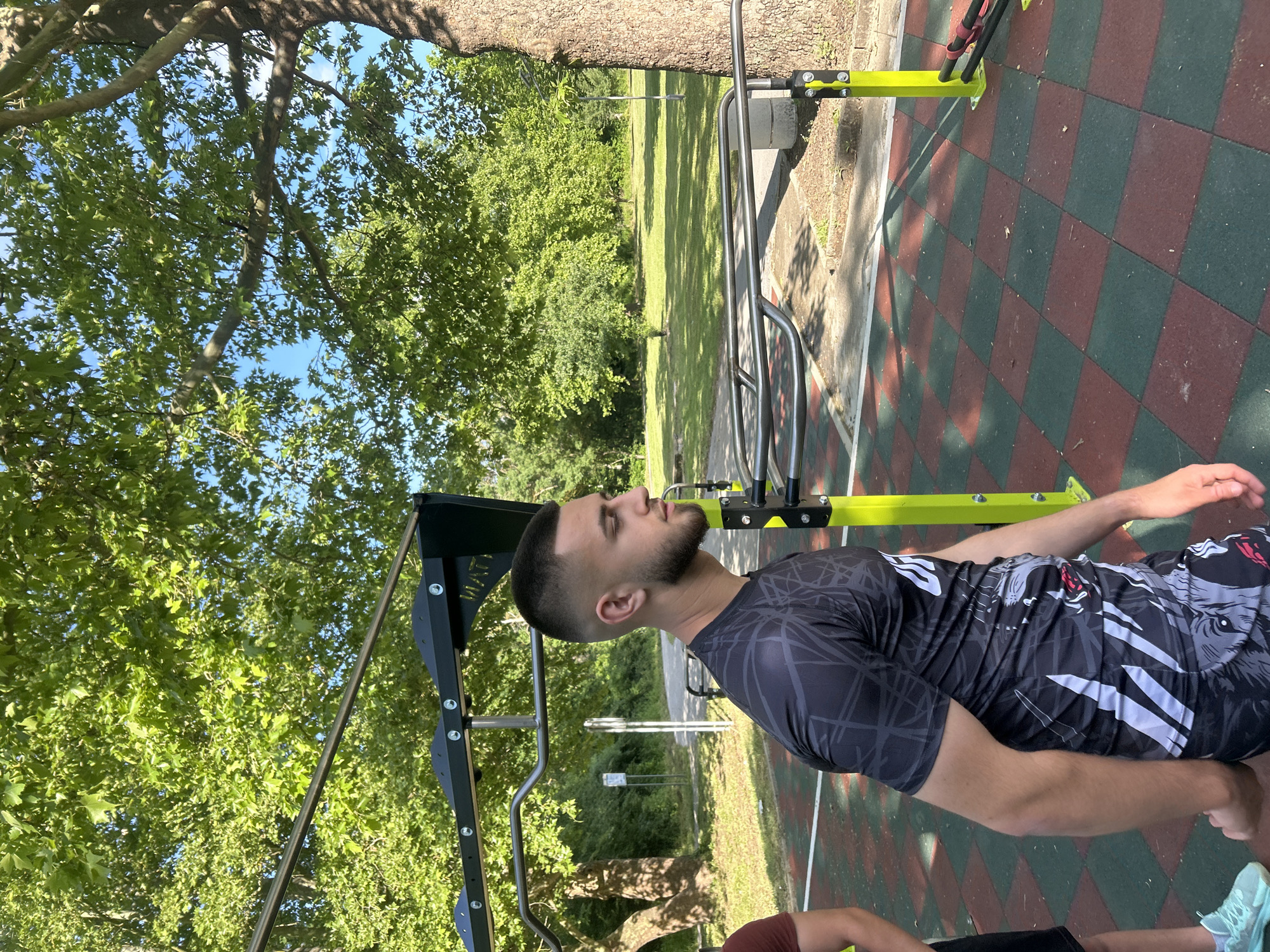 A man with a beard and short dark hair is seen in profile at an outdoor exercise area. He is wearing a gray and black patterned athletic shirt. The exercise area has green and red checkered flooring. There is a metal exercise structure with black and yellow components nearby. Trees are visible in the background. The location appears to be Elhovo, Bulgaria.