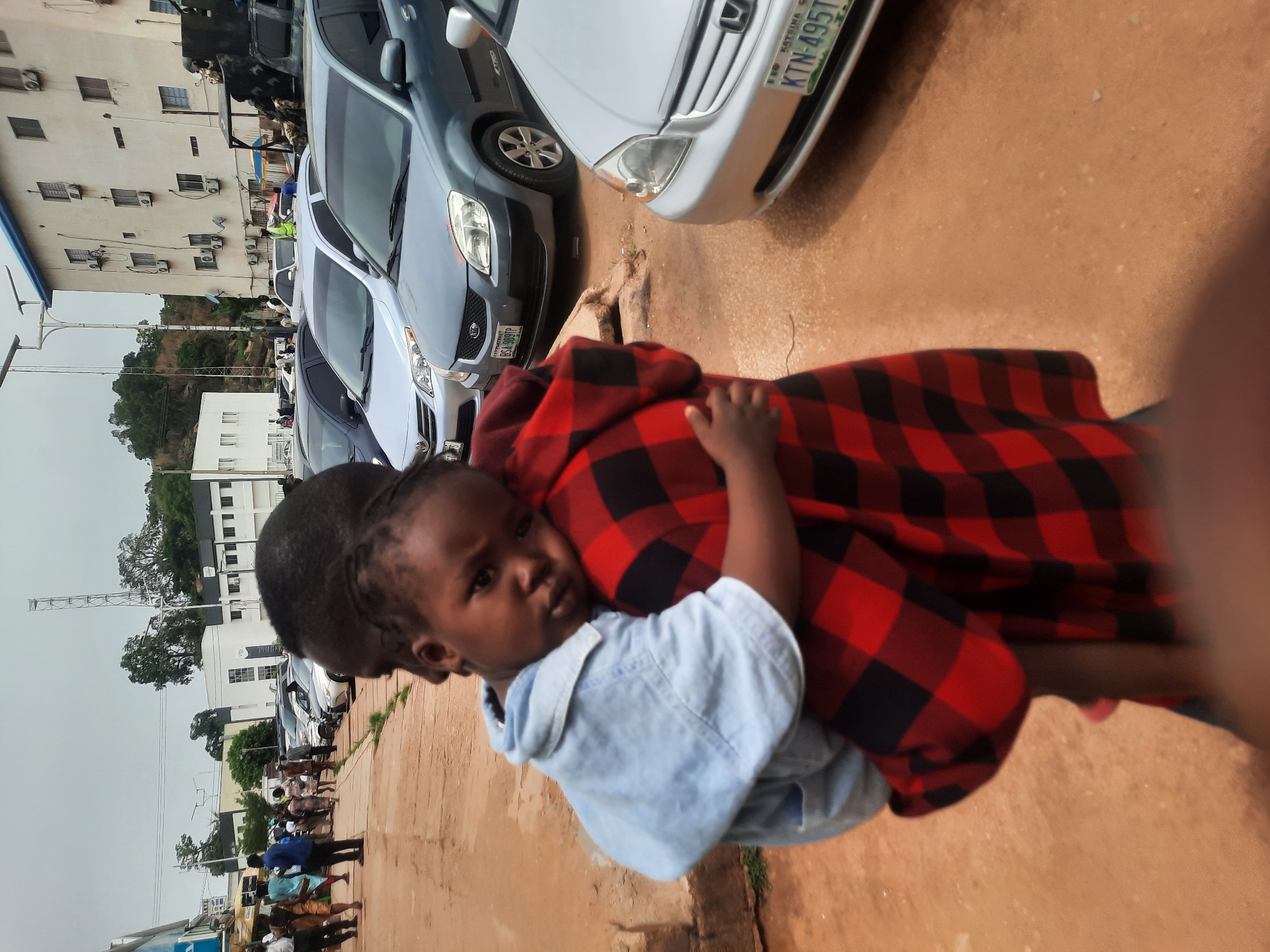 In Jos, Nigeria, an adult carries a young child in their arms. The child wears a light blue denim shirt. The adult is draped in a red and black checkered fabric. Cars line the street in the background. A group of people stand on a sidewalk in the distance. Buildings and trees are also visible.