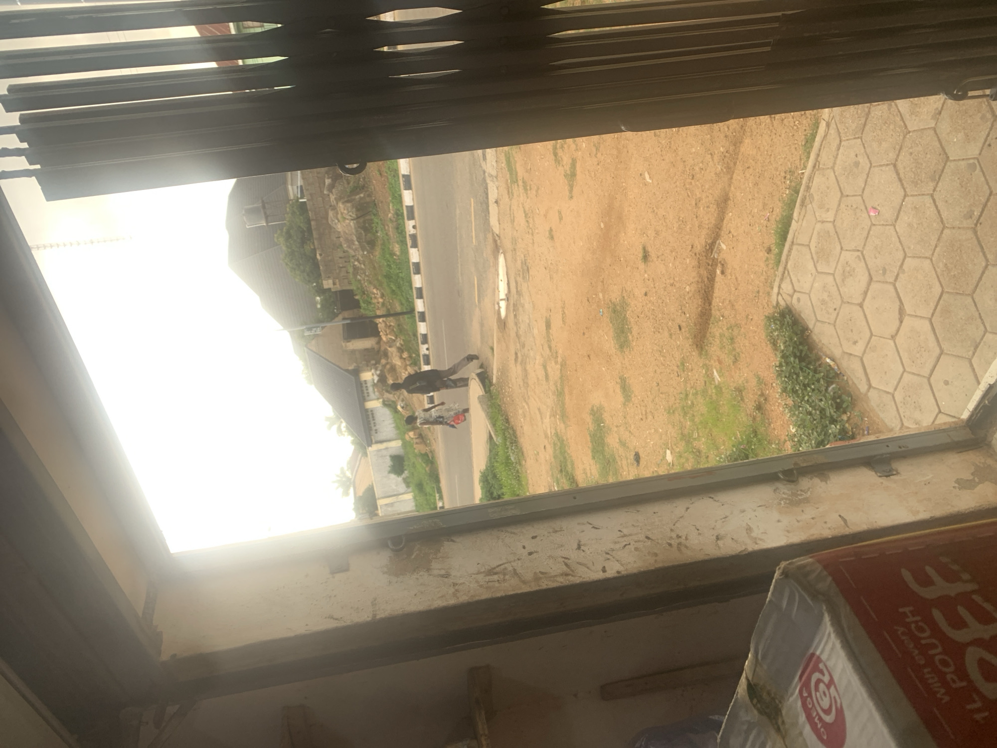 The image shows a view through a metal gate of a street scene outside. In the foreground, inside what appears to be a shop or covered area, there is a cardboard box with red and white packaging. Text on the box is partially visible, including "PEE" and "1 L POUCH With every". There is also a circular logo with the word "OMEGA" inside.
Outside, a road is visible with a striped white and black curb. Two people are walking on the road, one in a dark outfit and the other in a lighter, possibly red, outfit. The ground outside is mostly dry, sandy earth with patches of green grass. Buildings with stone and concrete construction are visible in the background, along with some trees. The sky is very bright, suggesting it is daytime, likely with sunny weather. The overall scene appears to be in a somewhat dusty, possibly rural or suburban area, consistent with a location in Jos, Nigeria.