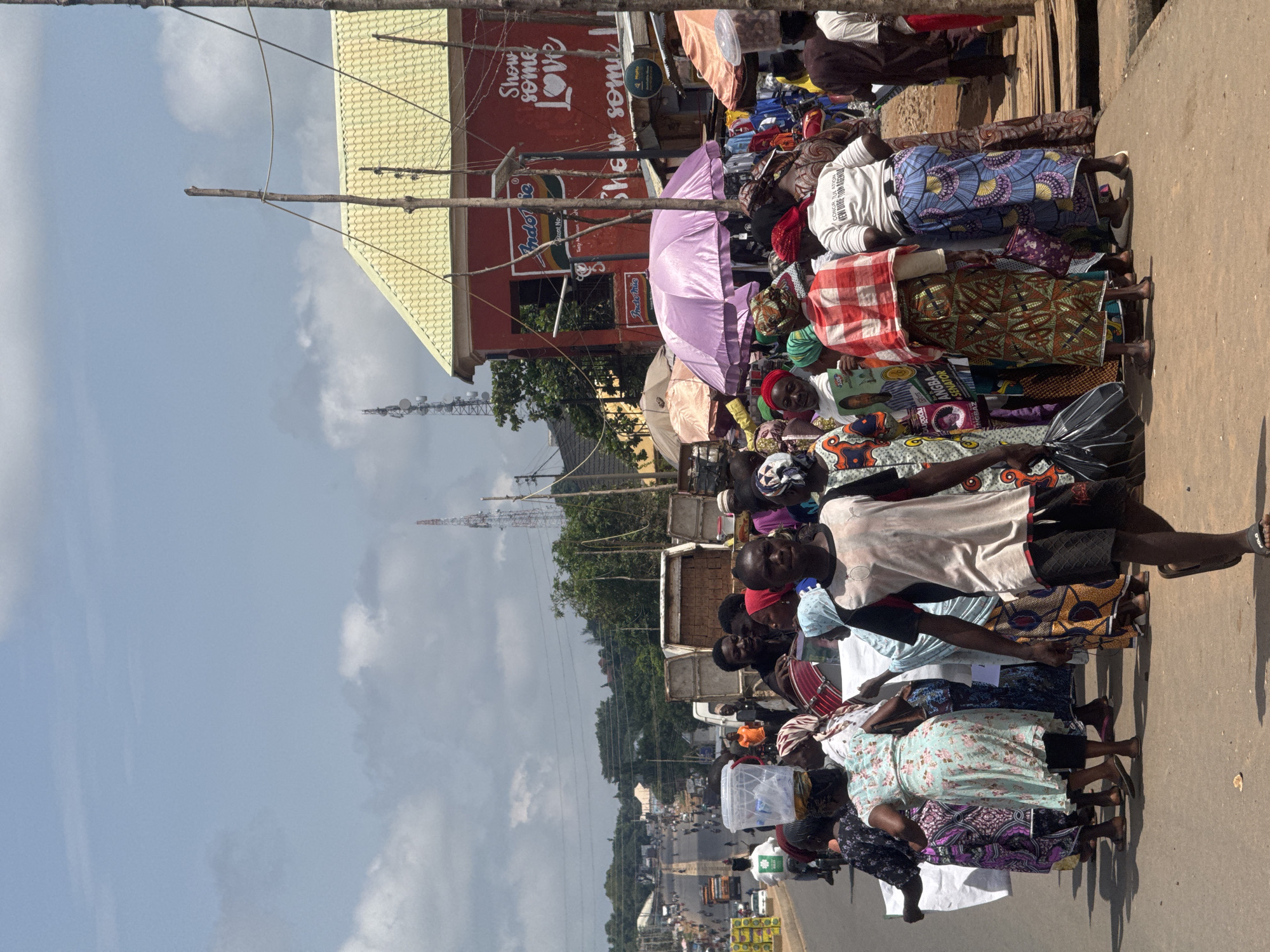 In Akwanga, Nigeria, a crowded street scene unfolds under a bright sky with scattered clouds. The foreground features a dense gathering of people, many dressed in colorful, traditional Nigerian attire. A young man in a white and black t-shirt and dark shorts walks towards the viewer, his face turned slightly away. To his right, a woman in a light blue floral dress carries a large clear plastic container filled with water on her head. She is surrounded by other individuals, some holding white sheets or bags.
In the mid-ground, a red building with a yellow tiled roof is visible, adorned with signs. One sign features the "Indomie" logo, and another painted message reads "Show some Love." Adjacent to this building, stalls are set up with clothing and other goods displayed. Several large umbrellas, including a prominent purple one, provide shade. A white truck with open-top containers filled with what appears to be bricks is parked on the side of the road.
The background shows a long, busy street with multiple lanes of traffic, including trucks and cars. Buildings and trees line the sides of the road, and a tall communication tower rises in the distance. The overall atmosphere suggests a bustling market or public gathering on a warm day.