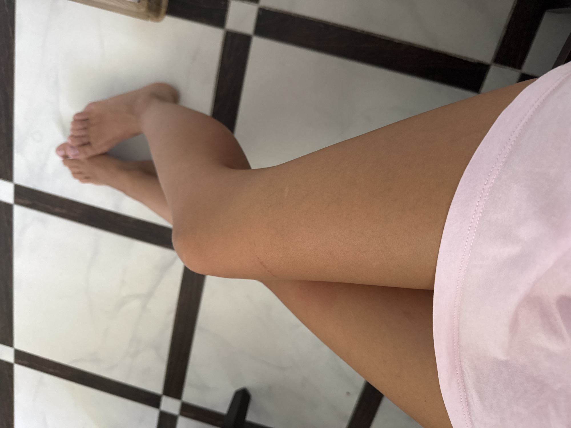 The image shows a person's legs and feet from a high-angle shot. The person is wearing a light pink garment that covers their midsection. Their legs are crossed at the knees, and their bare feet are visible. The floor is tiled with a black and white checkerboard pattern. There is no indication of the location being in Chișinău, Moldova, or any specific city.