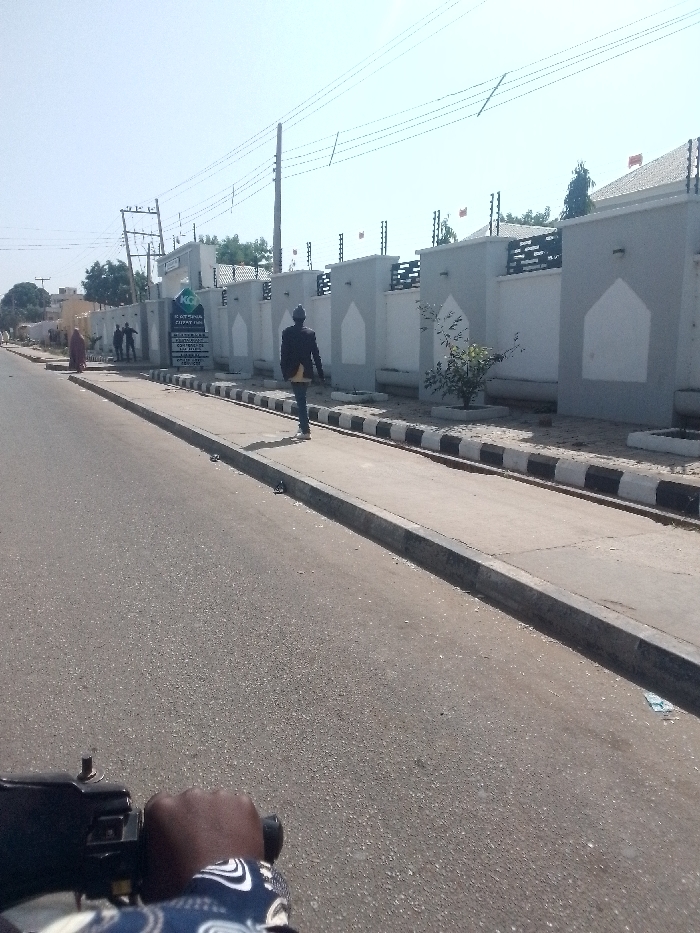 A street view presents a man in a dark jacket and light pants walking on a sidewalk bordered by a black and white curb. Behind him is a white wall with gray architectural accents and an electric fence. In the distance, other pedestrians are visible near a sign. A person's hand gripping handlebars suggests the photo was taken from a moving vehicle on the street. Power lines stretch across the sky.