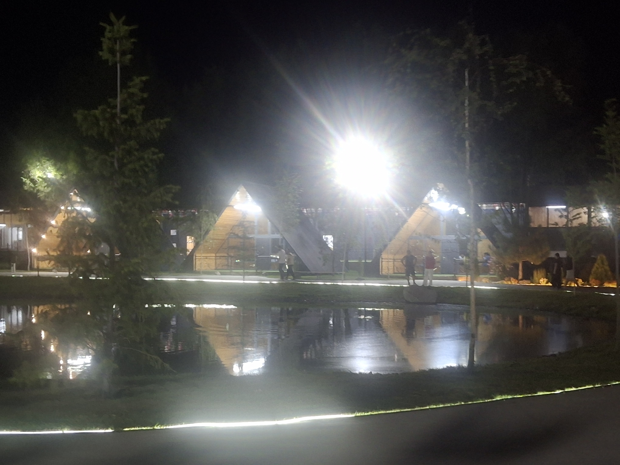 The image depicts a nighttime scene in what is described as the city of Aleksandrovka, Kyrgyzstan. In the foreground, a body of water reflects the lights from the surrounding area. The water's surface is smooth, suggesting a calm environment.
Along the far bank of the water, several triangular-shaped structures, possibly tents or small buildings, are visible. These structures are illuminated from within, casting a warm glow. The architecture appears modern and somewhat abstract. In front of these structures, a grassy area is visible, lined with low-profile lighting that creates a bright, continuous line along the edge.
Several individuals are present near the triangular structures. Two figures are standing together, appearing to converse. Another individual stands further to the right, silhouetted against the light. On the far right of the image, a group of people are gathered, some standing and others potentially interacting.
Dominating the upper portion of the image is a very bright light source, possibly a powerful spotlight or a very bright lamp, which creates a significant lens flare effect, obscuring some details in that area. Trees, some appearing to be evergreens, are interspersed throughout the scene, their dark silhouettes contrasting with the illuminated structures and the night sky. The overall atmosphere is festive and somewhat surreal due to the lighting and the unusual architectural forms.