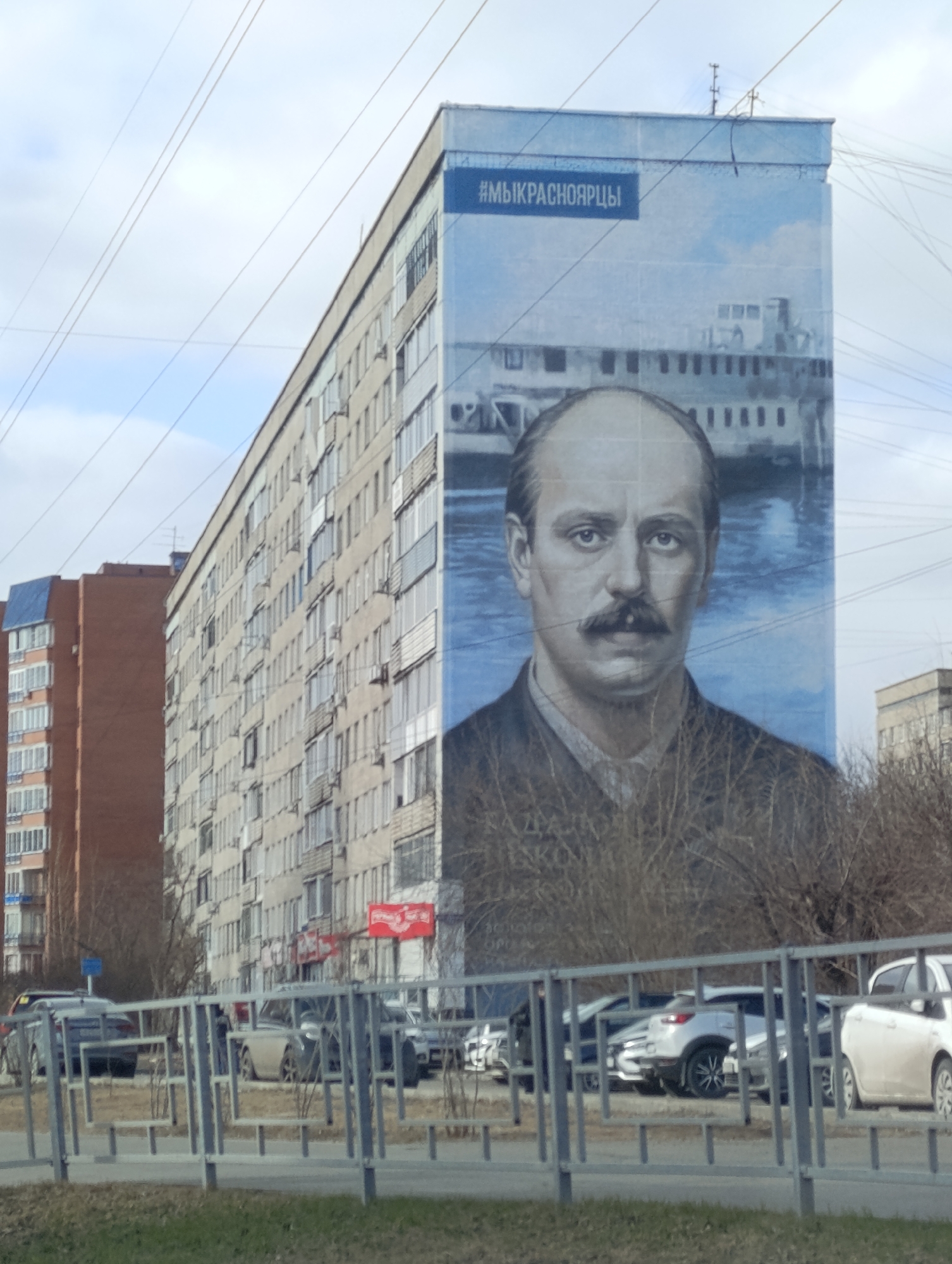 The image displays an urban scene in Krasnoyarsk, Russia, featuring a large mural on the side of a multi-story apartment building. The mural depicts a monochrome portrait of a man with a mustache and a serious expression, strongly resembling Anton Chekhov. Behind him, a paddle steamer or similar riverboat is visible on what appears to be a body of water, under a light blue sky. A blue banner across the top of the mural reads "#МЫКРАСНОЯРЦЫ" (meaning "#WEAREKRASNOYARSKCITIZENS"), confirming the location. Faint text below the portrait is unreadable but likely identifies the subject.
The apartment building itself is a typical Soviet-era or post-Soviet block, long and grey with numerous windows and balconies. To the left, another, taller brick residential building is partially visible. In the foreground, a metal fence separates the viewer from a street where several cars are parked. Bare trees or bushes, indicative of late winter or early spring, are scattered along the roadside. The sky is overcast, suggesting daytime with diffused light. The overall scene captures a residential area with an artistic public display.