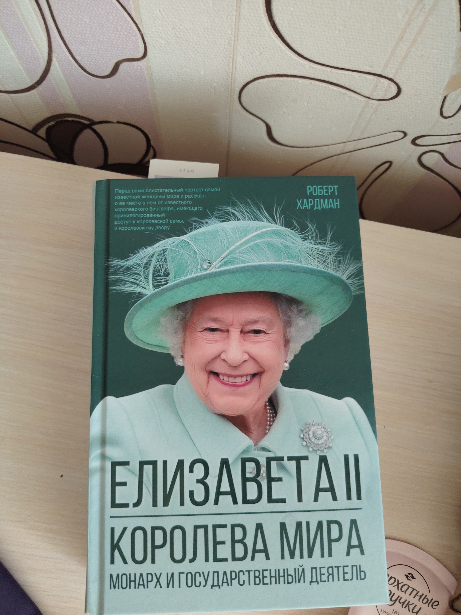 The image shows the cover of a book titled "ELIZABETH II QUEEN OF THE WORLD" in Russian. The book features a portrait of Queen Elizabeth II wearing a light blue hat with feathers and a pearl necklace. The background of the book cover is a dark green color. The book is placed on a wooden surface, likely a table. To the right of the book, a pink container with text and a woman's face is partially visible. The overall scene appears to be an indoor setting.