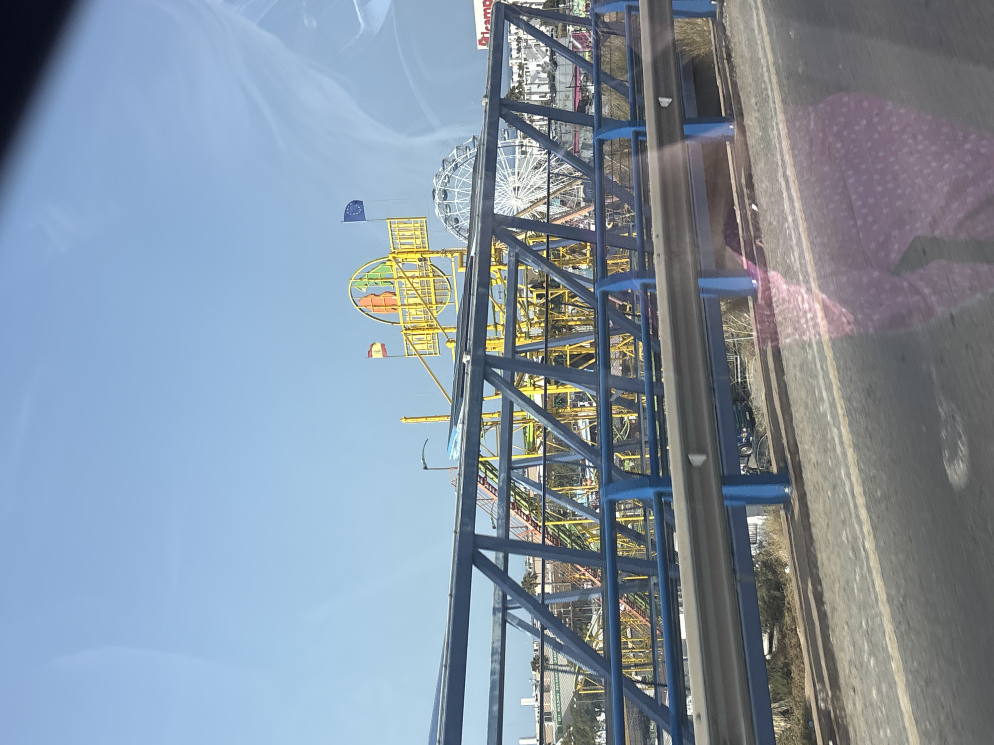 An eye-level, medium shot captures a view through a blue and grey metal pedestrian bridge. Beyond the bridge, a bright yellow and green roller coaster structure dominates the background, with red and orange tracks visible. To the right of the roller coaster, a white Ferris wheel stands tall against a clear blue sky. Two flags, one Spanish and one European Union, are also visible, fluttering in the breeze. In the distance, a cluster of buildings suggests an urban or semi-urban environment. The road in the foreground shows a yellow line marking the edge of the lane. A reflection of a person in a pink, polka-dotted garment is visible on the glass surface of the vehicle from which the photo was taken. The scene appears to be an amusement park or fairground, likely located in or near Orihuela, Spain, as indicated by the Spanish flag.