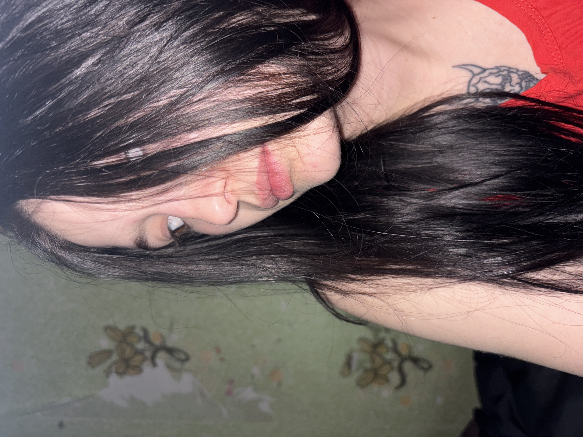 An indoor portrait, likely taken in Фоки, Russia, depicts a female subject from the chest up. Her head is turned slightly to the left, with her brown eyes directed toward the upper left, partially obscured by her long, dark, straight hair. Her lips are slightly parted, revealing no teeth. She is wearing a plain red top. A dark tattoo, resembling a horned skull, is visible on her upper right chest near the collarbone. The background consists of a light green wall covered with faded, patterned wallpaper featuring a repeating motif of yellowish-brown floral clusters. The wallpaper shows signs of wear and discoloration. A dark, indistinct object is visible in the lower left portion of the frame.