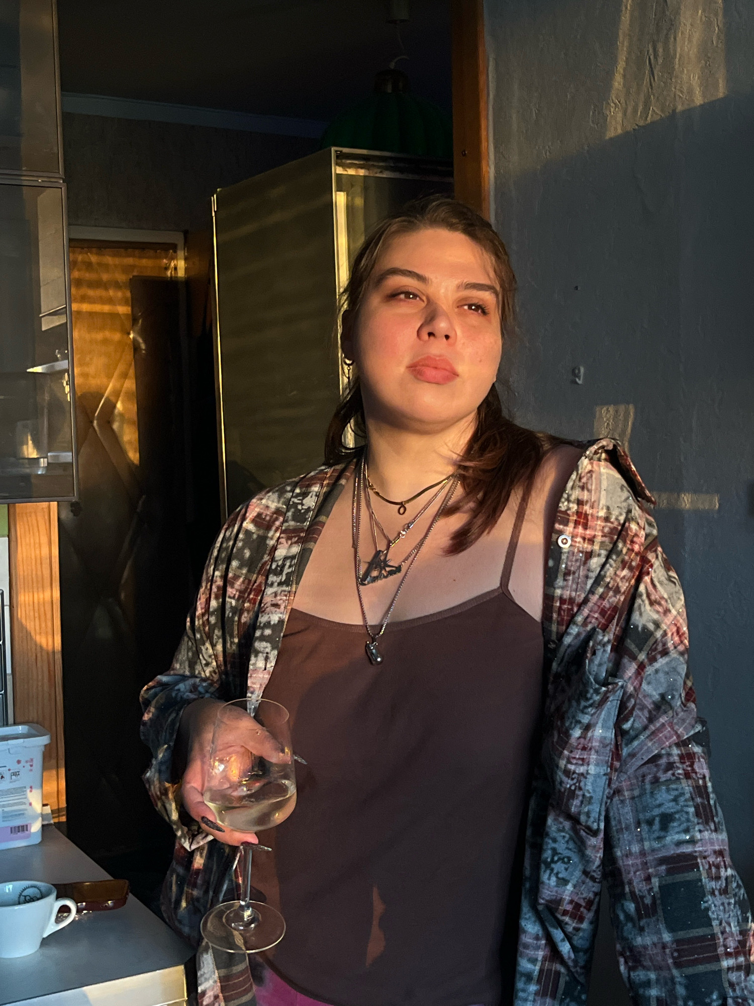 A woman stands in a dimly lit indoor space, likely a kitchen or utility area. She is looking upwards and to the right, with her head tilted slightly. She is holding a wine glass in her right hand, which contains a pale liquid. She is wearing a brown camisole and an unbuttoned plaid shirt over it. She has several necklaces on, including one with a prominent pendant.
The background is cluttered with various objects. To the left, there are shelves with containers, and a white mug is visible on a counter. Behind her, a large metallic appliance, possibly a refrigerator or industrial equipment, is present. The lighting in the scene is dramatic, with warm sunlight casting long shadows on the wall behind her, suggesting it might be early morning or late afternoon. The overall atmosphere is one of casualness, possibly a moment of reflection or relaxation. The location is not specified to be Moscow, Russia.