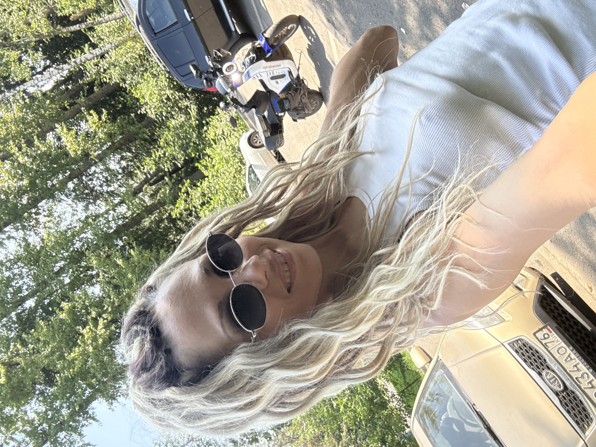 A woman with blonde dreadlocks and sunglasses smiles at the camera, taking a selfie outdoors. She is wearing a white tank top. Behind her, a blue and white scooter is parked, and two cars are visible: a gold Kia to her left and a dark SUV to her right. Trees and foliage form a natural backdrop. The scene appears to be a street in a residential area, possibly Rusanovo, Russia, given the license plate on the Kia.