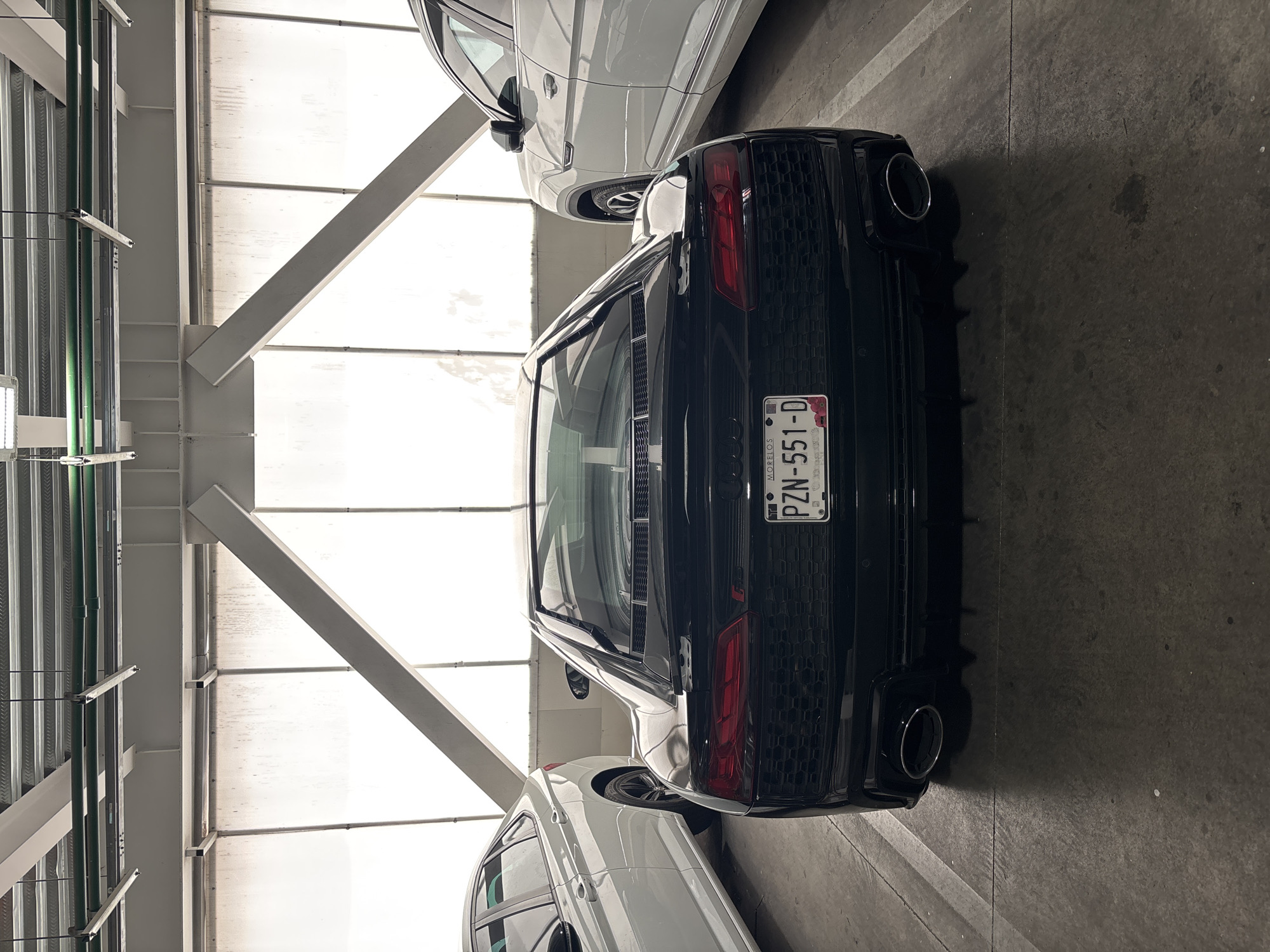 The image depicts the rear view of a black Audi R8 parked in a parking garage. The license plate is visible, reading "PZN-551-D" and includes the state of Morelos logo, which does not match the location of San Bernardino Tlaxcalancingo, Mexico. A white vehicle is partially visible on either side of the R8. The parking garage has a bright, skylit roof supported by metal beams. The floor is concrete. There are no people visible.