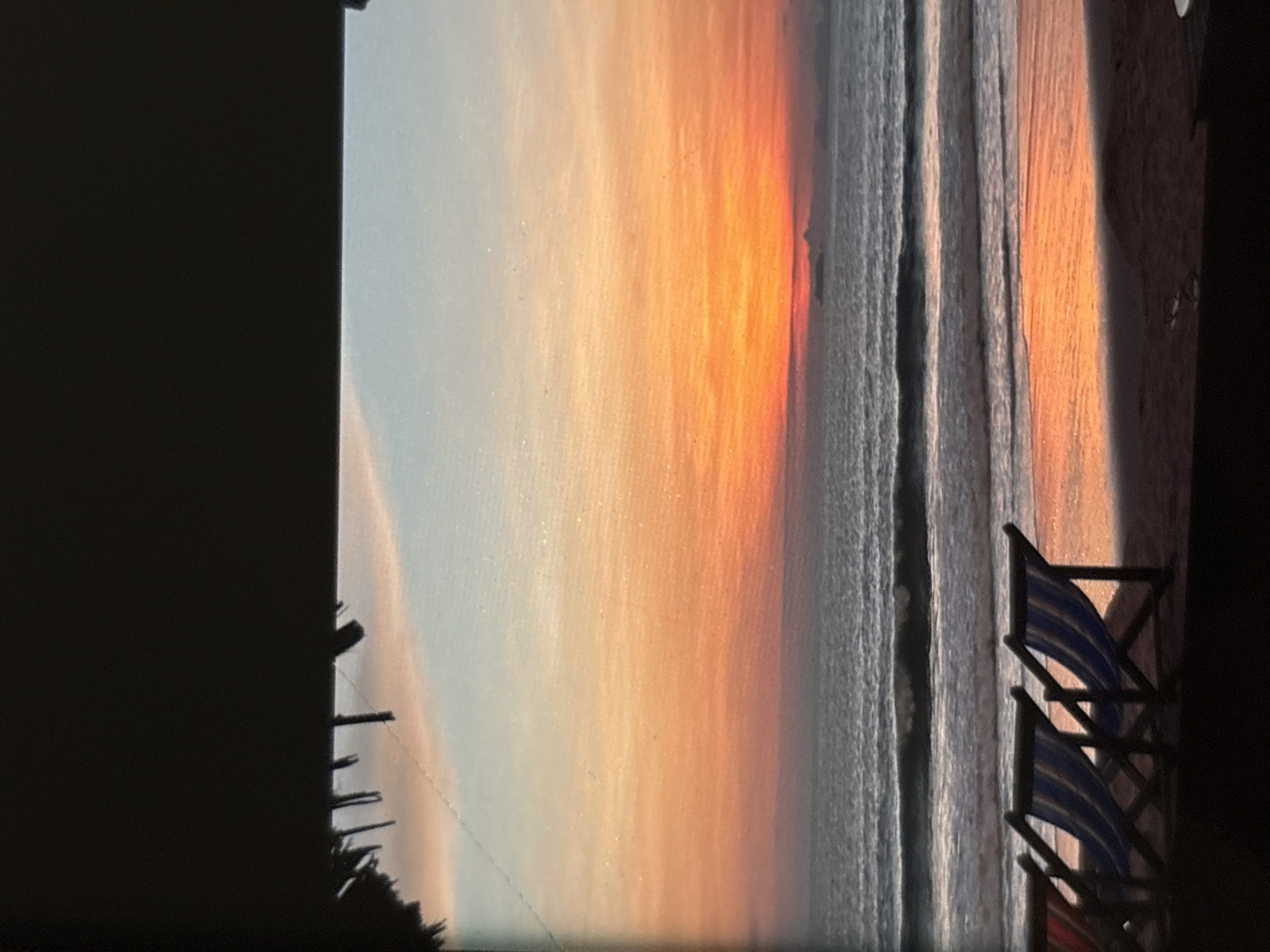 This image depicts a tranquil beach scene at sunset, likely in Chalong, Thailand. In the foreground, three beach chairs with striped blue and white canvas are positioned on the sand, angled towards the ocean. The chairs are empty. The ocean is calm, with gentle waves rolling onto the shore. The water reflects the vibrant colors of the sunset, creating an orange and pink glow on the wet sand. In the distance, the horizon is a soft blend of orange, pink, and blue, characteristic of a sunset. A faint silhouette of land or islands can be seen on the horizon. The top of the image is framed by a dark, shadowy area, suggesting the viewpoint is from under a shelter or overhang, possibly a thatched roof structure, with some frayed edges visible.
