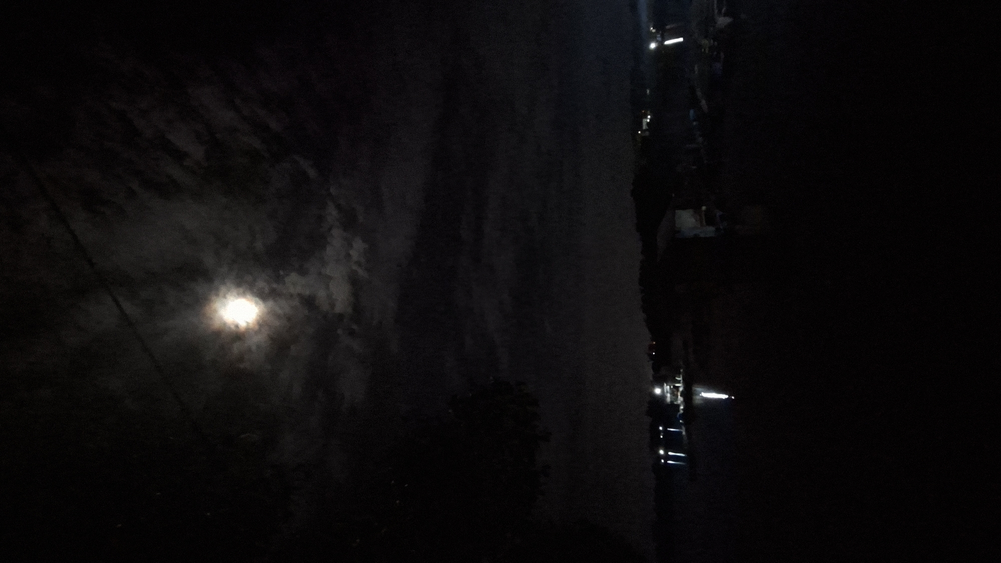 The image captures a nighttime scene in Lhokseumawe, Indonesia, dominated by darkness and the glow of the moon. The moon, a bright orb, is partially obscured by clouds, casting an ethereal light on the cloudy sky.
In the foreground, a silhouetted tree is barely discernible against the dark background. A power line runs diagonally across the left side of the frame, adding a man-made element to the natural landscape.
To the right of the frame, the scene shifts to a more urban or populated area. A row of lights suggests buildings or structures, possibly along a waterfront or street. Some of these lights appear to be streetlights or lights from vehicles, illuminating a pathway or road. Further into the darkness, a few more distinct lights are visible, possibly indicating a cluster of buildings or a waterfront development.
The overall mood is one of quiet darkness, with the moon providing the primary illumination. The image appears to have been taken on a cloudy night, as evidenced by the diffusion of the moonlight. The presence of artificial lights on the right suggests a blend of natural and urban environments. No discernible text is visible in the image.