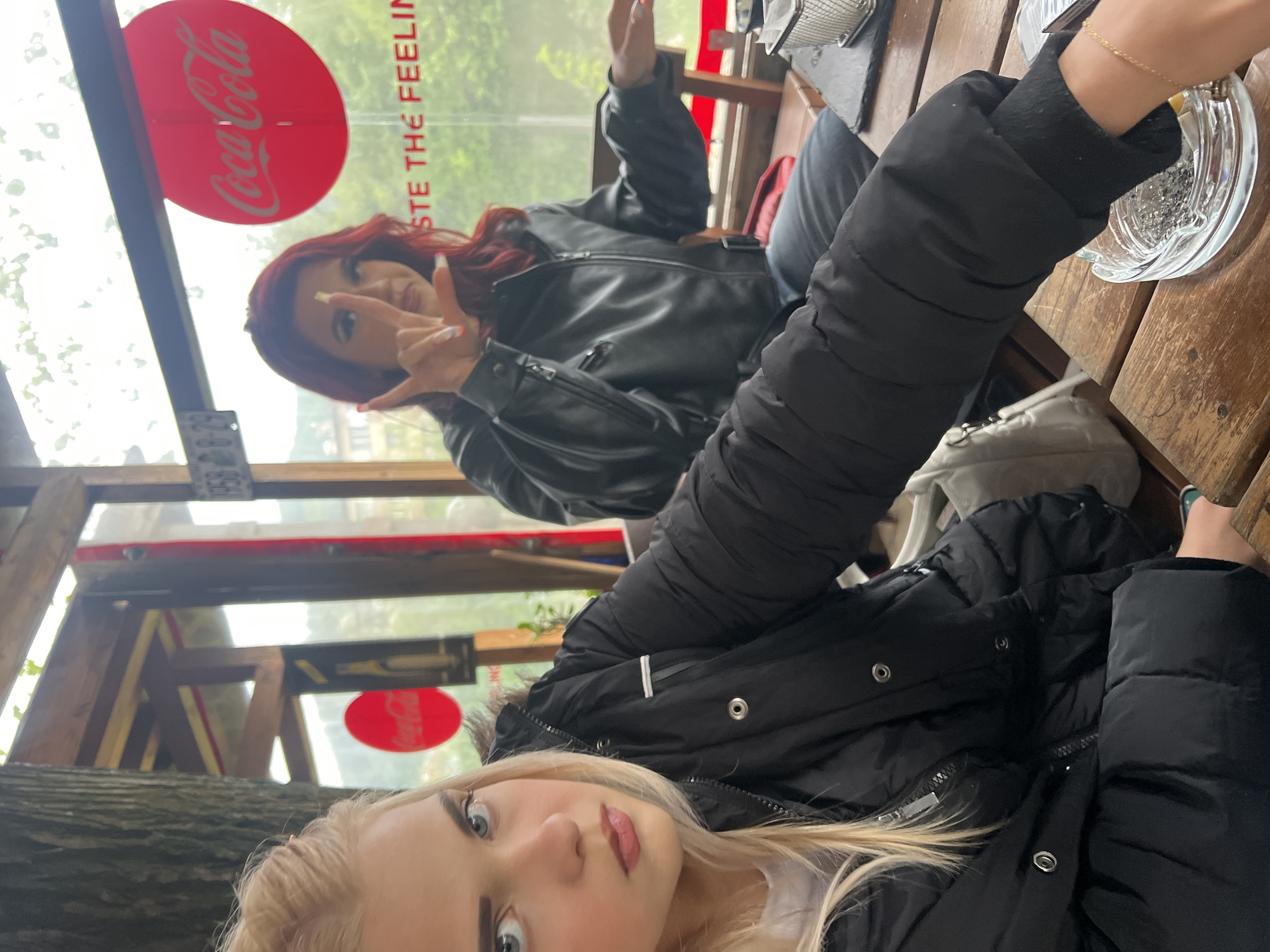 Two women are seated at a wooden table in an outdoor setting. One woman with red hair is wearing a black leather jacket and making a hand gesture. The other woman, with blonde hair, is wearing a black puffy jacket. An ashtray is on the table. A Coca-Cola sign is visible in the background. The location is identified as a city of Unknown, Unknown.