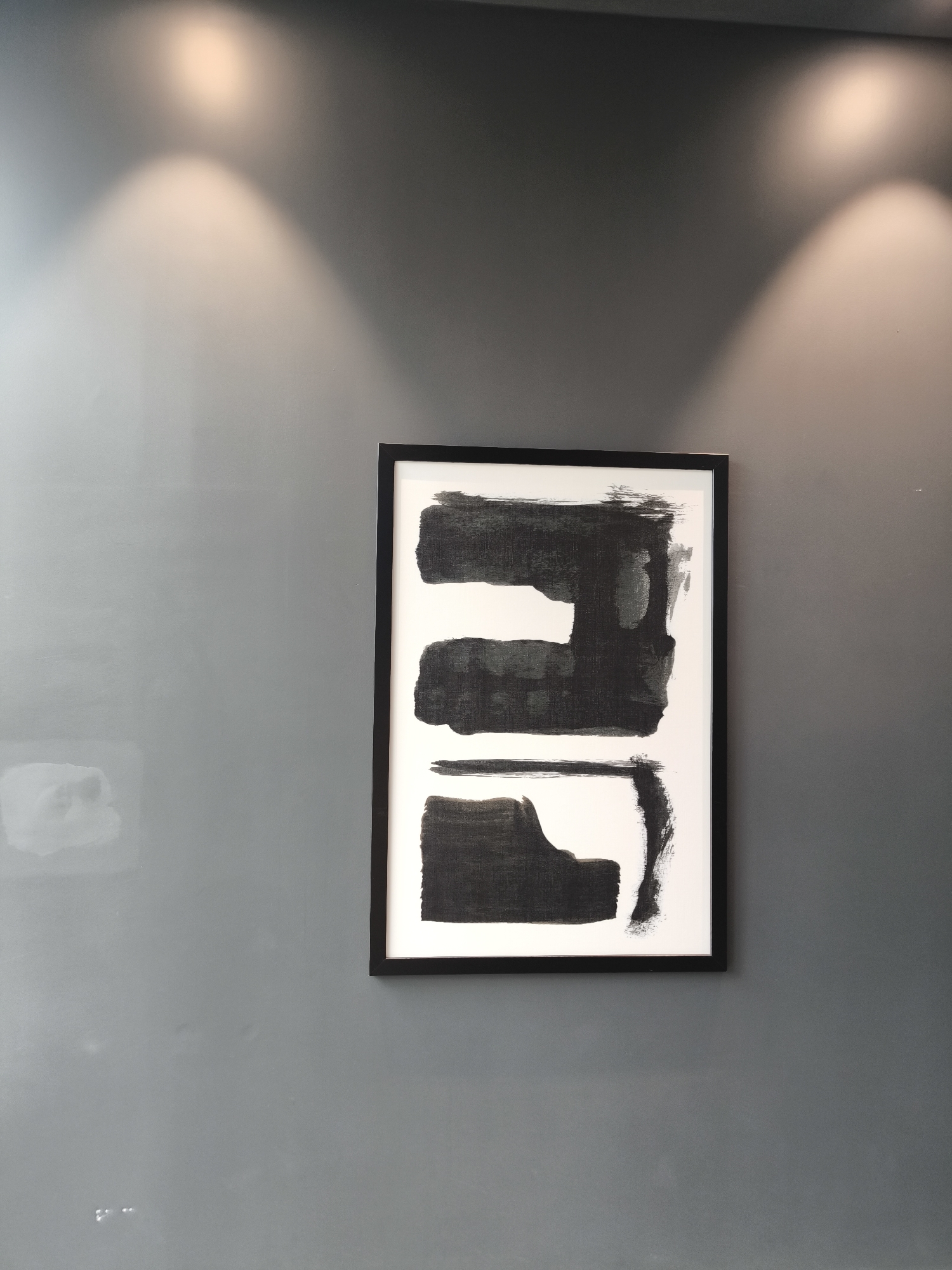 The image shows a framed abstract painting with black brushstrokes on a white background, hanging on a smooth, dark gray wall. Two spotlights illuminate the wall from above, creating pools of light above and to the sides of the painting. The painting is centrally located in the frame, oriented vertically, and features several distinct black shapes, some solid and some with visible brushstroke texture. The wall exhibits a subtle texture and appears to be a matte finish. There are no people or other objects visible in the scene. The overall impression is minimalist and modern, likely an interior space. The location of this scene in Gatineau, Canada is not discernible from the provided image.
