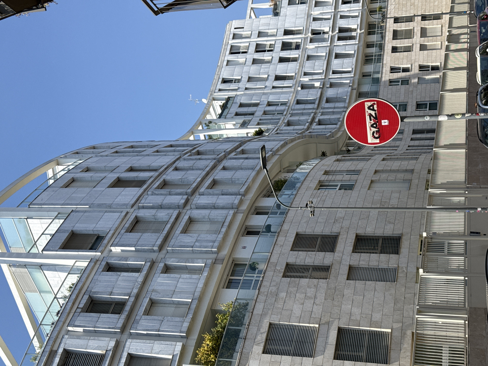 The image shows a view of a curved, multi-story building in Milan, Italy. The building has a light-colored facade with rows of windows. The architecture is modern with curved features. A red and white "No Entry" sign with the word "GAZA" written on it is attached to a pole in the foreground. The sky is blue.