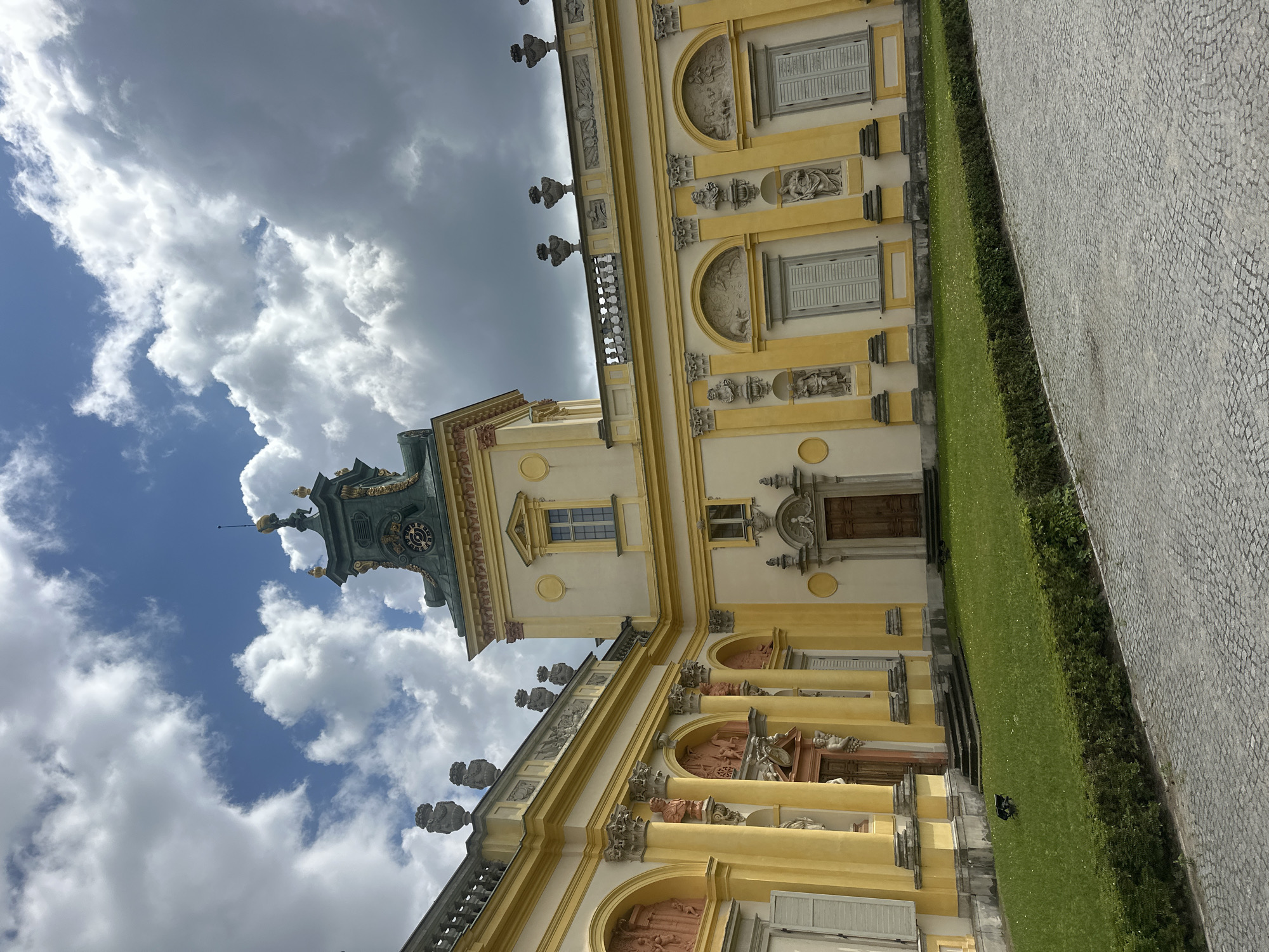 The image shows the Royal Palace of Wilanów in Warsaw, Poland. The palace is a Baroque-style residence located in the Wilanów district. The scene is captured from an exterior perspective, showcasing a section of the yellow-colored facade adorned with columns, arched niches, and decorative sculptures. The building features a prominent clock tower with a green roof and gold accents. A well-maintained green lawn with scattered white flowers and a trimmed hedge runs in front of the palace. The foreground is paved with cobblestones. The sky above is a mix of blue and dramatic, cloudy formations. There are no people visible in the image.