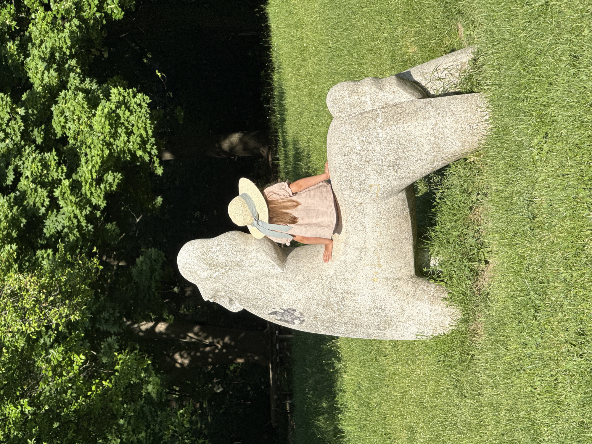 A young person wearing a straw hat with a blue ribbon and a light-colored dress is nestled within a large, weathered, gray stone sculpture. The sculpture is situated on a grassy lawn, with lush green trees and foliage in the background. The scene appears to be outdoors on a sunny day. The subject is not actively engaged with the sculpture, rather seated within it. The location is Sofia, Bulgaria, as this sculpture is part of the collection at the Borisova Gradina park.