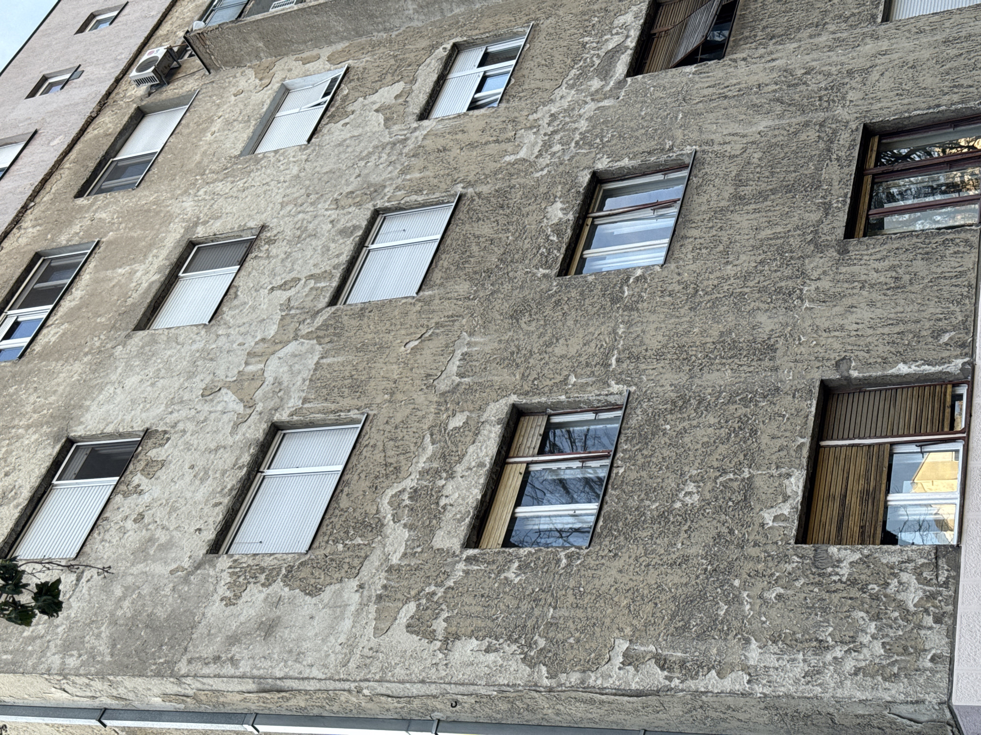 The image shows the exterior facade of an old building in Belgrade, Serbia. The building is made of concrete and is several stories high. The facade is weathered and shows signs of deterioration, including peeling paint. There are numerous windows along the facade. Some of the windows have blinds or wooden shutters. The windows vary in their state of repair; some are open, and others are closed. An air conditioning unit is affixed to the building near the top left. The perspective of the photo is from a low angle, looking up at the building. The sky is not visible. The overall scene depicts urban decay and neglect.