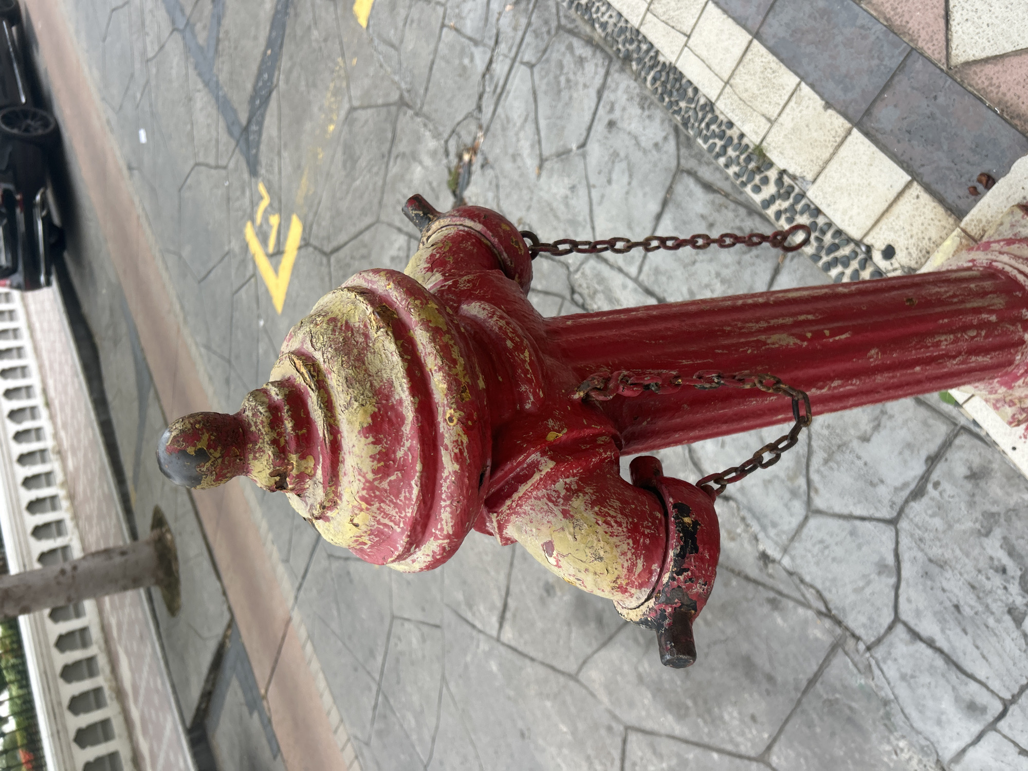A close-up, eye-level shot shows a red fire hydrant with chipped yellow paint on a textured, grey paved street in George Town, Malaysia. The hydrant has two spigots, each with a rusty chain attached, one chain hanging down to the left, the other to the right. The top of the hydrant has a ribbed, dome-like cap. In the background, a black car is parked on the street, and a white decorative fence lines the sidewalk, beyond which is a tree trunk. The pavement is a mosaic of irregular grey stone tiles, with a strip of smaller, dark grey pebbles and white tiles running along the right edge of the frame. Yellow arrows indicating traffic direction are visible on the pavement in the middle ground.