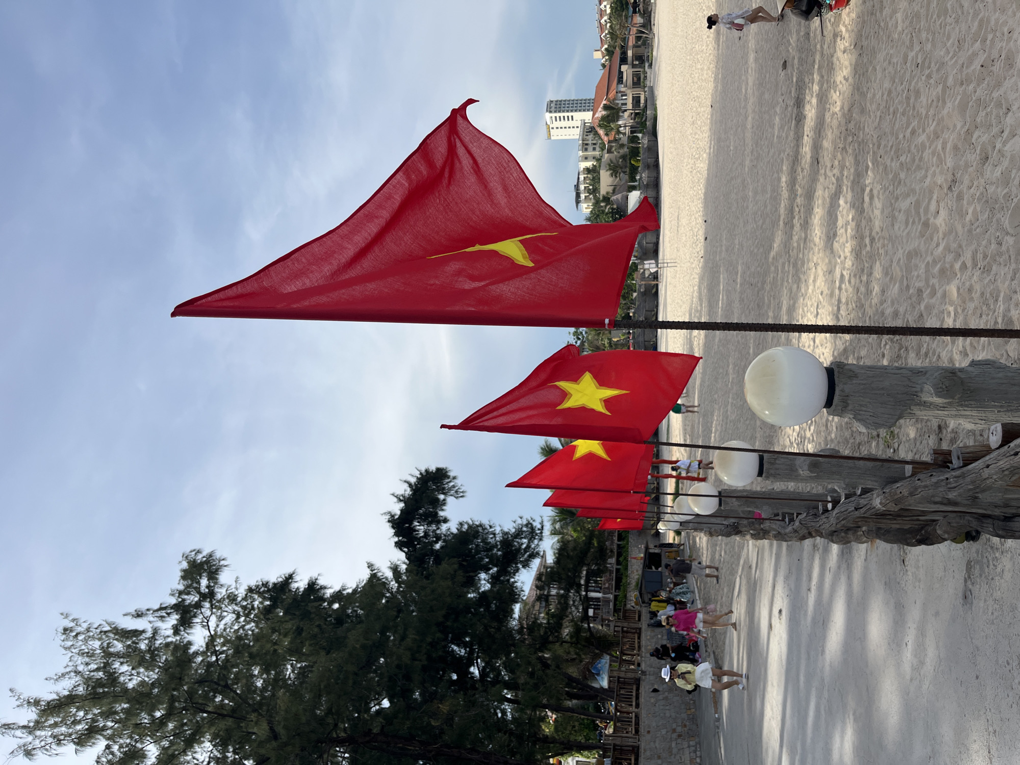 A line of red Vietnamese flags with yellow stars are planted along a sandy beach, marking a pathway on the left side of the frame. The flags are mounted on dark poles and some are catching the breeze, causing them to billow. Behind the flags, the sand stretches out to the ocean, which is not visible. In the background, there are buildings, palm trees, and a tall hotel structure. Several people are walking along the pathway and on the beach, some dressed casually in summer clothing, including women wearing hats and light outfits. The pathway is made of concrete and bordered by a rustic wooden fence with spherical lamps mounted on its posts. Trees with dense foliage are visible on the far left of the image, casting shadows on the pathway. The sky is a light blue with some scattered clouds. The overall scene depicts a coastal area, likely a tourist spot, in Phu Thuy Ward, Vietnam.