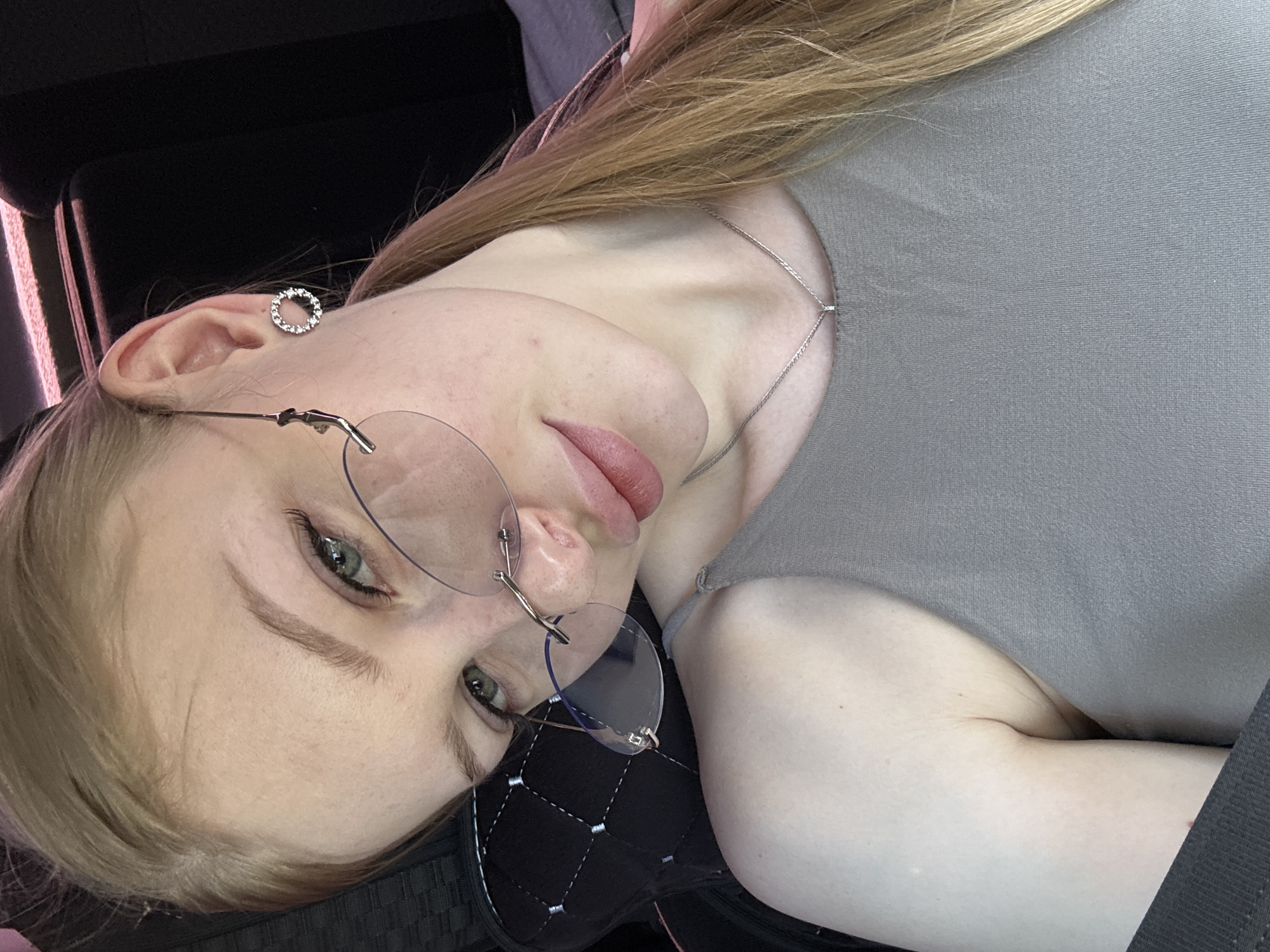 The image depicts a fair-skinned woman with light blonde hair wearing round glasses. She has green eyes and is wearing a silver necklace and earring. She is wearing a gray top. The background is dark and appears to be inside a vehicle, possibly a car. The location in the city of Perm, Russia cannot be determined from the image.