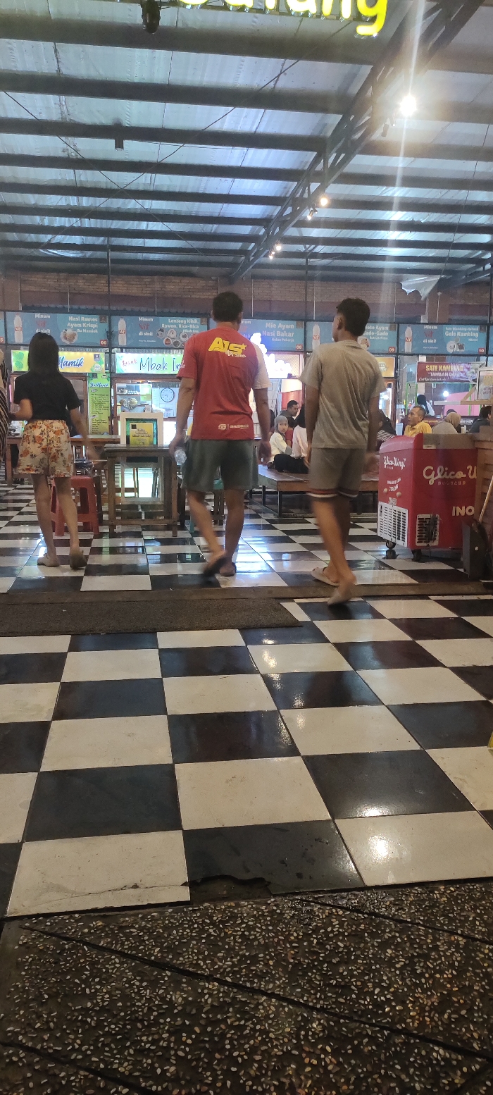The image shows a black and white checkered tiled floor in a food court in Mojokerto, Indonesia. The location is marked by the coordinates -7.458, 112.448. Three individuals are walking through the food court. One man is wearing a red shirt with the letters "AIS" on the front and dark blue shorts, another man is wearing a grey long-sleeved shirt and dark blue shorts, and a woman is wearing a floral patterned skirt and a dark-colored shirt. There are also several tables and chairs in the background, as well as a large red cooler with the words "Glice U" on it. There are signs on the walls with Indonesian language, and other individuals can be seen sitting at the tables. The scene is brightly lit, with fluorescent light fixtures visible in the ceiling. The food court is largely empty except for the three individuals walking through. The floor is slightly wet.