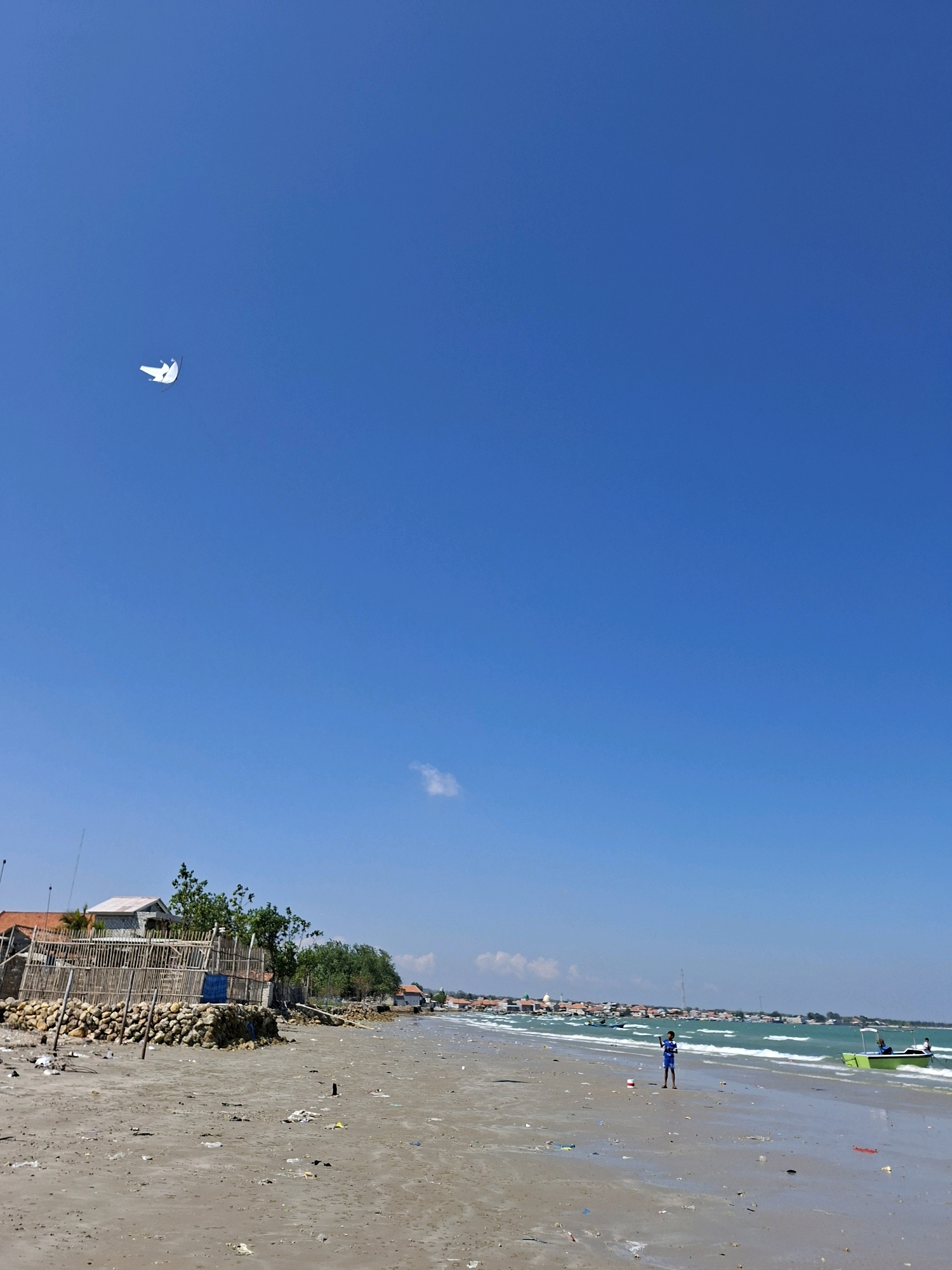 The image depicts a bright, sunny day on a beach in Camplong, Indonesia. The sky is a clear, vibrant blue, with a single, small white cloud visible in the upper right quadrant. In the upper left portion of the sky, a white kite in the shape of a bird is flying.
The beach itself is sandy and appears somewhat littered with various pieces of trash. The water is a turquoise green, with moderate waves rolling onto the shore. In the distance, a line of buildings and trees suggest a coastal town.
On the beach, a young person wearing a blue shirt and shorts stands near the water's edge, facing the sea. To the right, a small, bright green boat with two people in it is partially in the water. Further down the beach, closer to the viewer, there are structures made of wood and bamboo, possibly part of a fence or building under construction, with piles of rocks and more debris. The overall impression is a tropical beach scene, albeit one affected by litter.