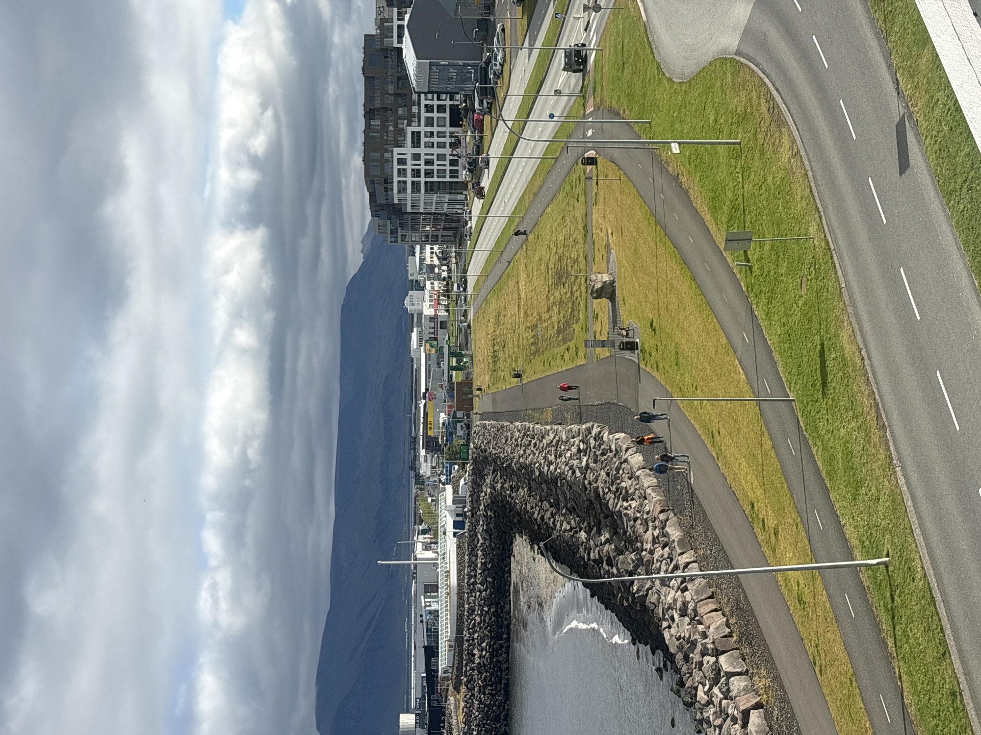 The image captures a coastal area in Reykjavik, Iceland, likely along the Sæbraut promenade. A wide, paved walkway runs alongside a rocky breakwater that protects a small sandy beach and the sea. The walkway is bordered by a green, grassy area with a separate, narrower path for cyclists or pedestrians.
Several individuals are seen walking along the main pathway. In the foreground, a group of four people are walking together, while further along the path, another group of three people can be seen. A single person on a bicycle or scooter is visible on the narrower path to the left of the road.
Beyond the walkway and grassy area, a multi-lane road curves through the scene, with several cars and a dark-colored SUV present. Parking areas are visible along the roadside, with numerous vehicles parked.
In the background, a cluster of modern buildings, including a large, multi-story building with a grid-like facade, line the cityscape. Further in the distance, industrial buildings and structures are visible, with a prominent white building and smokestacks. The backdrop is dominated by a range of dark, mountainous terrain under a sky filled with dramatic, gray clouds, with patches of blue sky visible. A single, distant object, possibly a plane, is noted in the sky. The lighting suggests it is daytime.