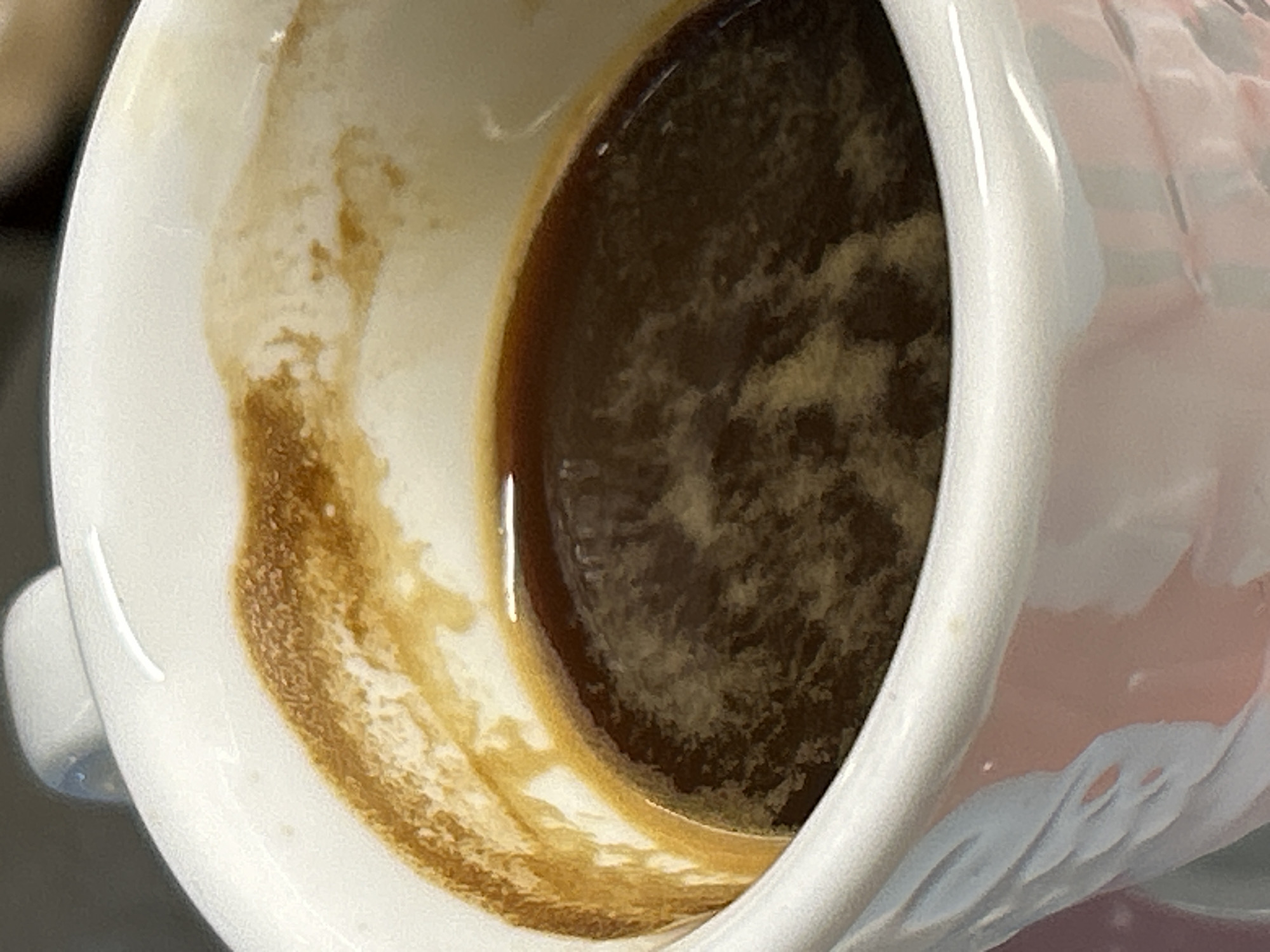 A close-up shot shows the inside of a white ceramic mug, largely empty. The mug contains a dark brown liquid with a lighter brown rim and sediment. Residue clings to the inside of the mug, indicating consumption of the liquid. The mug handle is visible on the left, and the exterior has a decorative pattern. The mug's pattern contains the colors white, pink, and light blue. The location and association with Burgas, Bulgaria, cannot be determined from the visual information alone.