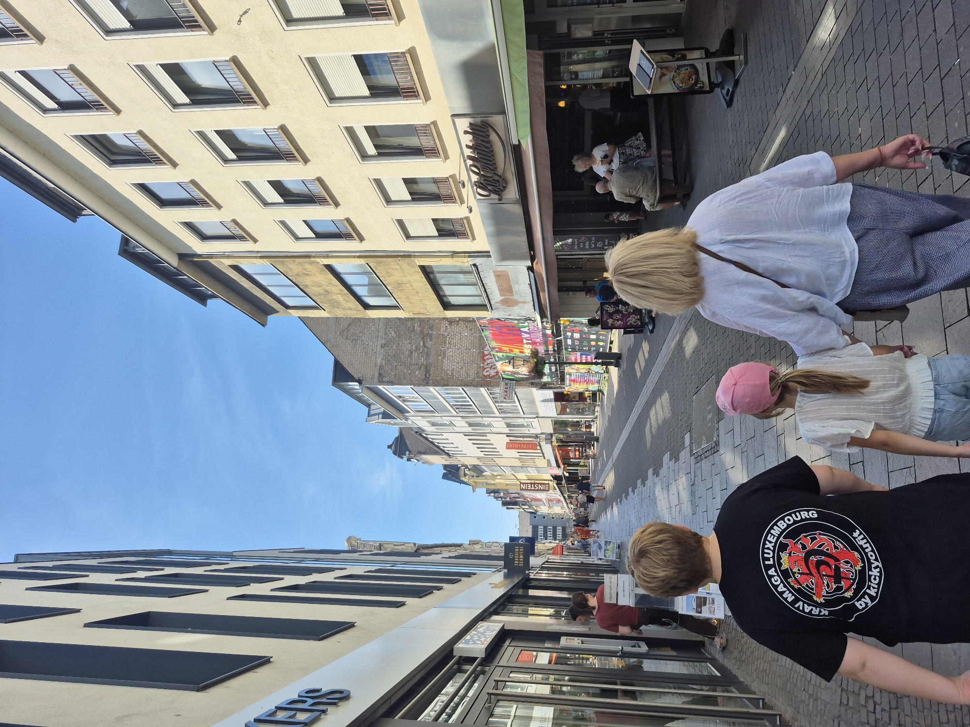 A group of people are walking down a paved pedestrian street lined with buildings in Koblenz, Germany. The buildings are multi-story structures with shops on the ground floor. A sign reading "La Gelateria" is visible above one of the shops. The sky is clear and blue. In the foreground, a man wearing a black t-shirt with a red and black logo is walking away from the camera. A young girl in a pink hat is walking beside him. A woman with blonde hair, wearing a white shirt and patterned pants, is walking slightly ahead of them. Several other pedestrians are visible in the background, walking in both directions. The street is paved with interlocking bricks.