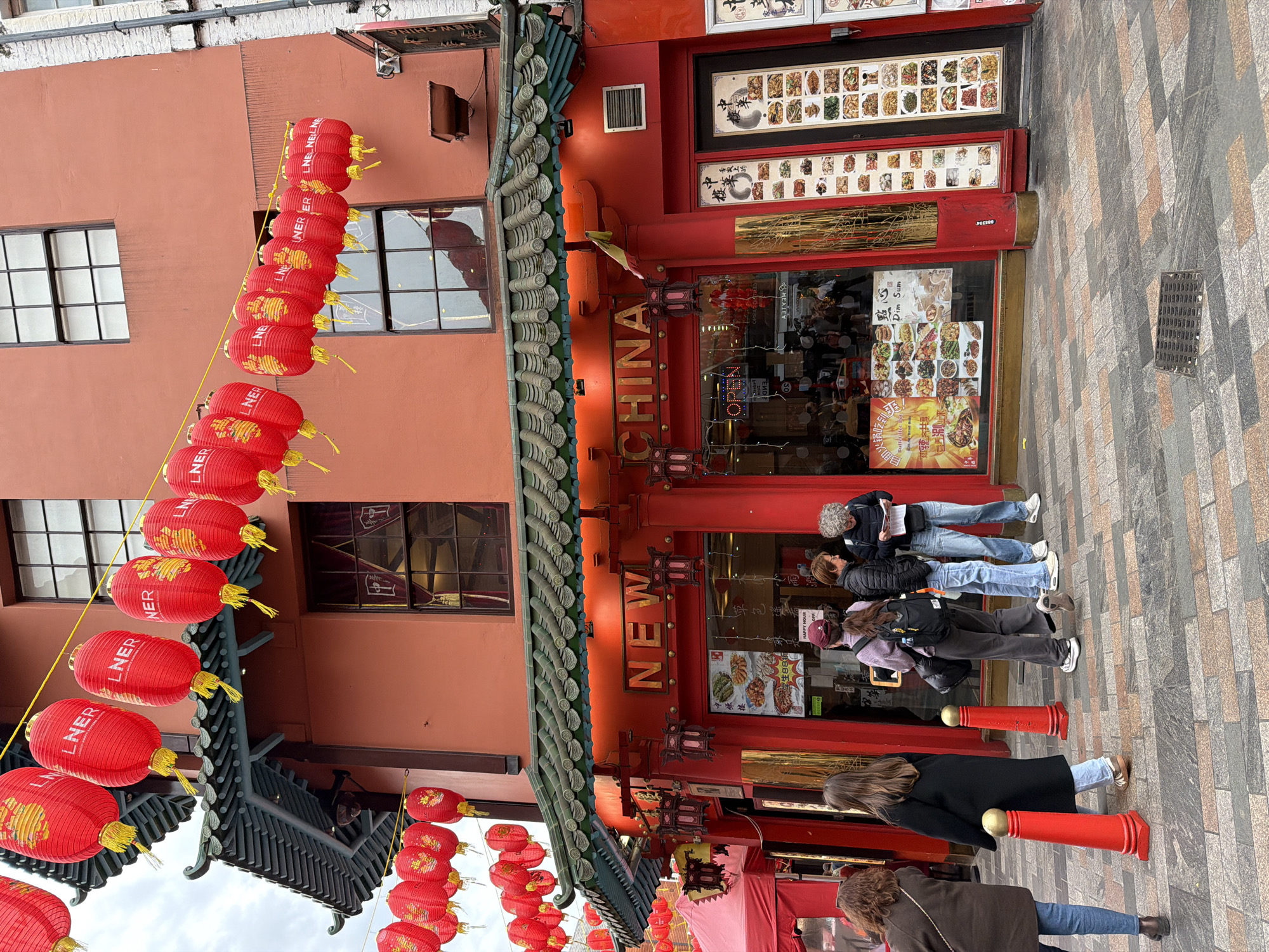 The image shows the facade of a restaurant in Chinatown, London, United Kingdom. The restaurant has a traditional Chinese architectural style, with a red facade, a green tiled roof, and red lanterns hanging outside. The name "NEW CHINA" is displayed prominently above the entrance. The restaurant's menu is displayed in the front windows. People are standing outside the restaurant. The street is paved with patterned stones.