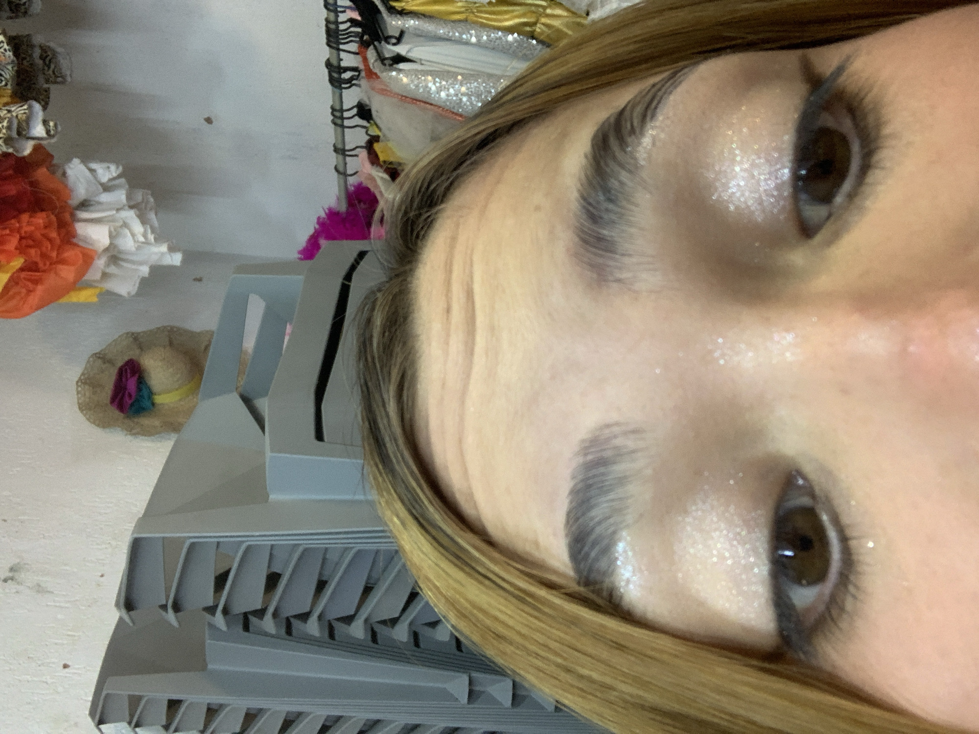 A close-up, tilted image shows a portion of a person's face, including their eyes, eyebrows, and forehead. The skin appears to have a light layer of glitter. The person has brown eyes and dark eyelashes. Strands of blonde hair frame the face.
In the background, there are stacks of gray plastic trays or containers. A straw hat decorated with colorful ribbons is visible. Parts of costumes with orange and white elements are also present.
The location is unspecified within the city of Porlamar, Venezuela.