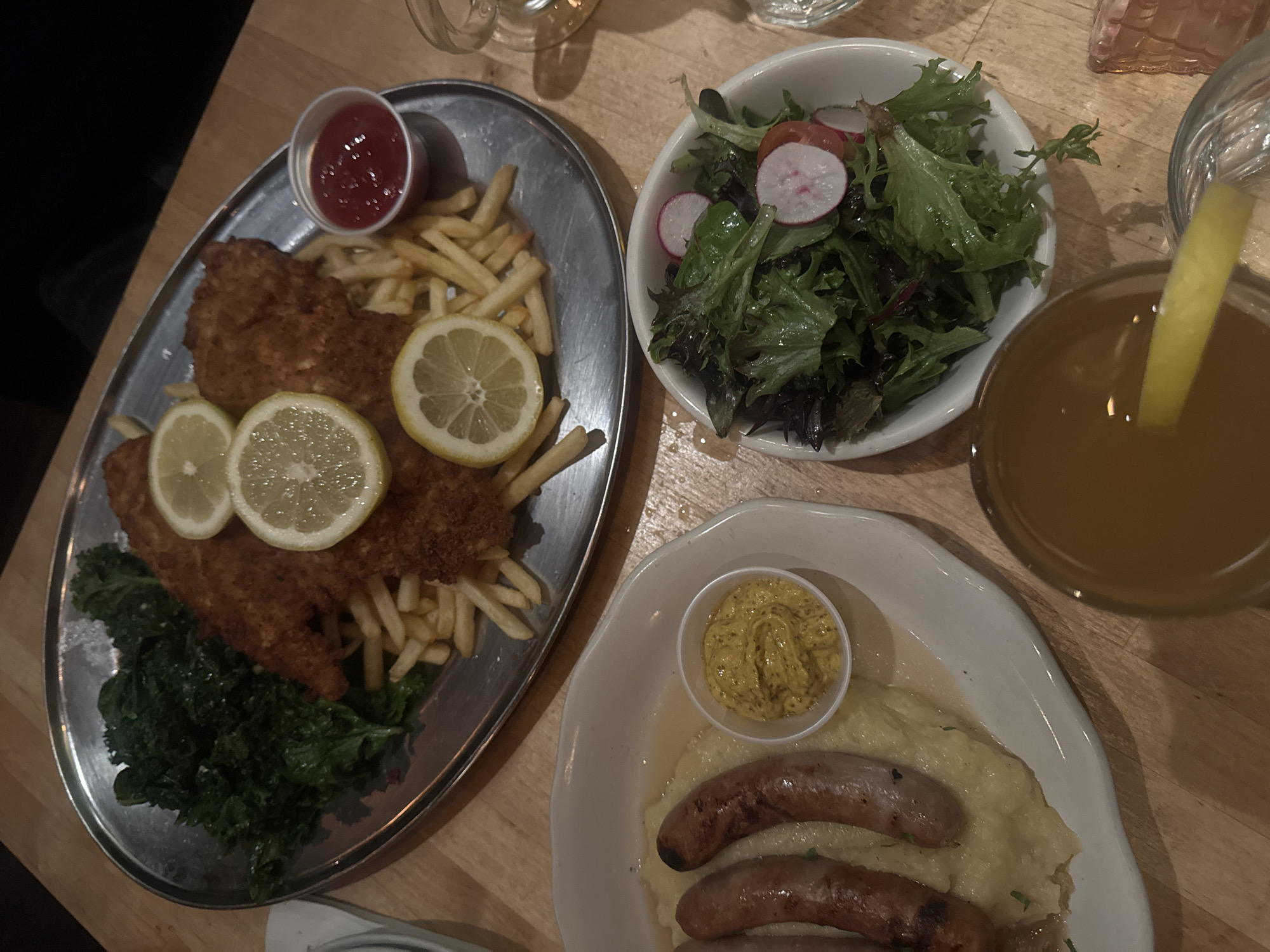 The image shows a tabletop restaurant scene with two plates of food and beverages. On the left, a silver plate holds what appears to be schnitzel with French fries, lemon slices, a small container of ketchup, and a serving of dark greens. To the right, a white plate contains sausages on a bed of mashed potatoes, with a small dish of mustard. A salad in a white bowl with radishes and mixed greens is above the sausage plate. To the right of the salad, a glass of amber-colored liquid with a lemon slice is visible. The table appears to be wooden. The location cannot be determined from the image.