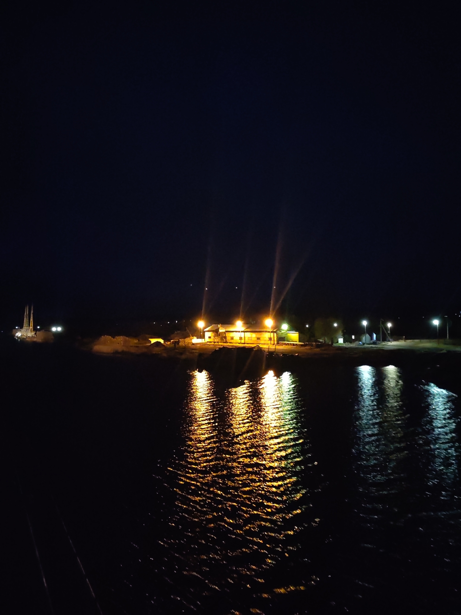 A night-time shot shows a body of water with lights reflecting off the surface. Across the water is a building with illuminated windows and lights above, casting beams into the dark sky. Streetlights dot the shoreline to the right. In the distance to the left, silhouettes of structures are visible. The location is Unknown, Unknown.