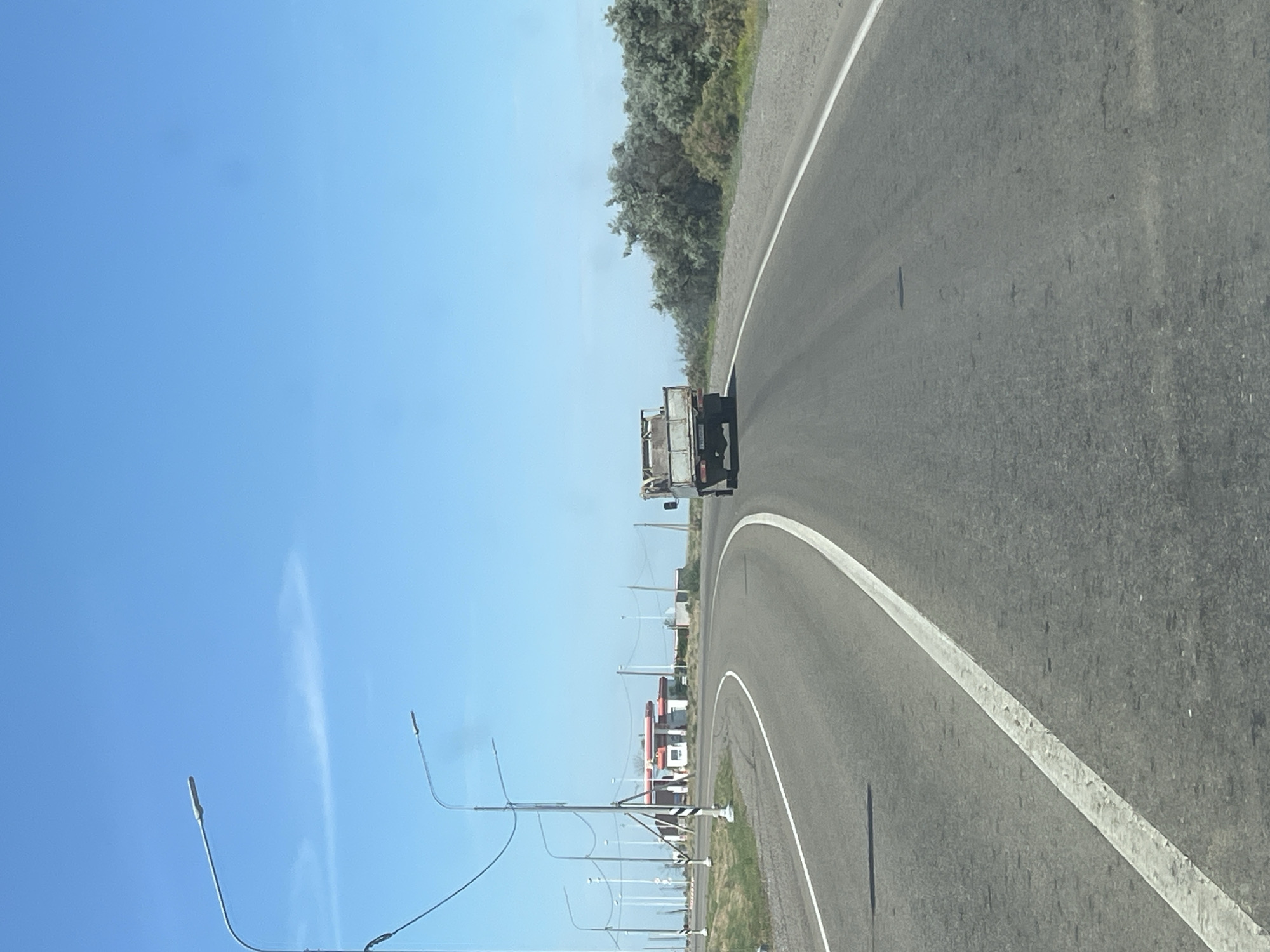 The image shows a clear blue sky with a few wispy clouds over a highway. A truck is driving away from the viewer on the right side of the road. The highway curves to the left, leading to a gas station with a red roof and white buildings in the distance. Streetlights line the left side of the road, and trees and bushes are visible on the right. The scene appears to be in a rural or semi-rural area. The location is identified as Bakhtemir, Russia.