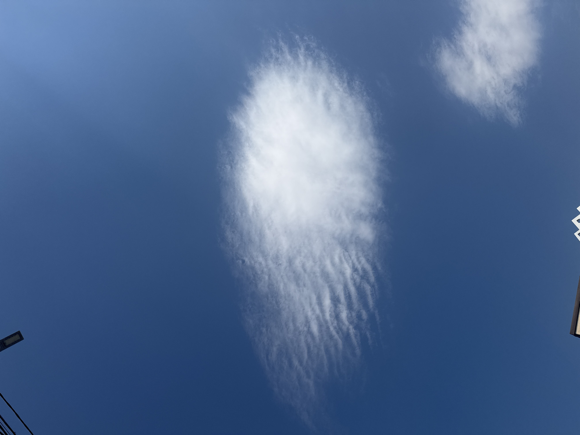 The image shows a bright blue sky with a large, wispy cloud formation in the center. The cloud has a textured appearance with delicate, wave-like patterns. To the upper left, a dark, elongated object, possibly a street lamp or utility pole, is visible against the sky. In the lower portion of the image, the top of a building with a dark roof is partially visible, along with a white decorative element. A smaller cloud formation is also present in the lower right corner. The scene appears to be an outdoor urban environment, consistent with Bishkek City, Kyrgyzstan, given the clear sky and typical urban infrastructure.