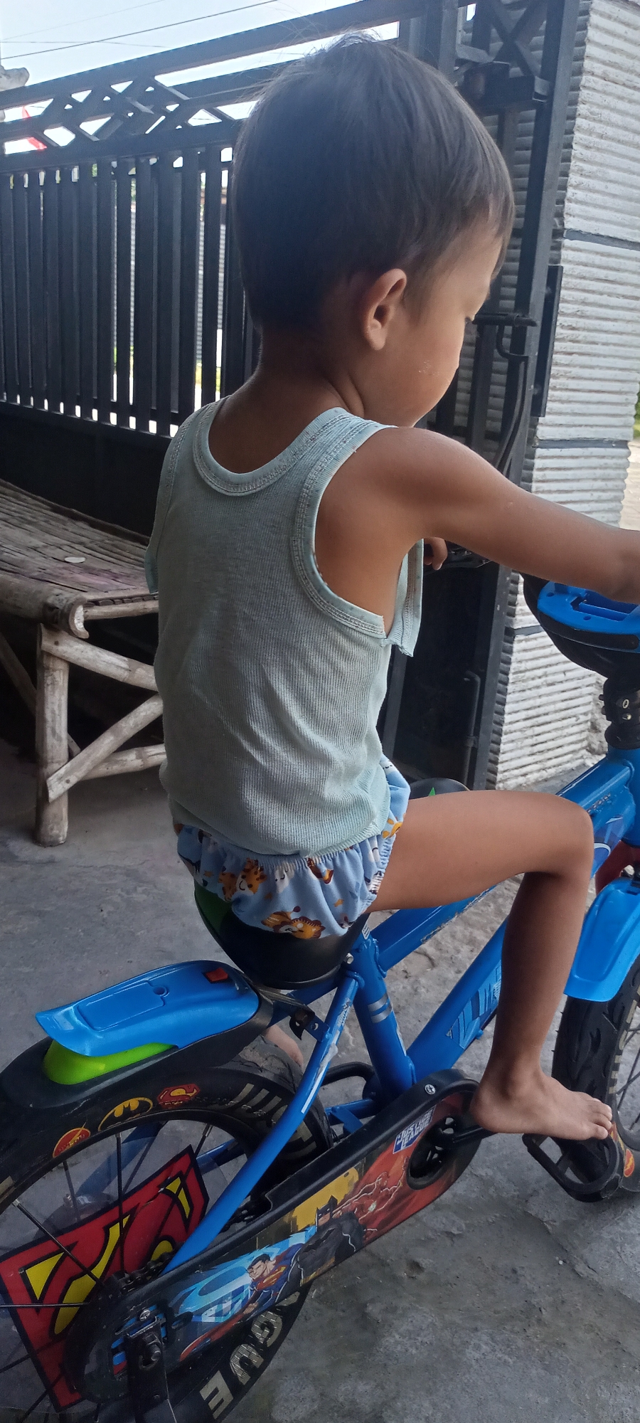 A young child, facing away from the camera and to the right, is seated on a blue bicycle. The child is wearing a light blue tank top and blue shorts adorned with a cartoon animal print. Their bare feet are visible, with one resting on a pedal. The bicycle features a blue seat and handlebars, and a decorative chain guard with images of superheroes like Superman and Batman. The scene appears to be outdoors, on a concrete surface, with a dark metal fence and a wooden bench visible in the background. The setting is likely within a residential area of Gempolklutuk, Indonesia, suggested by the architectural elements and outdoor environment. The lighting indicates it is daytime.