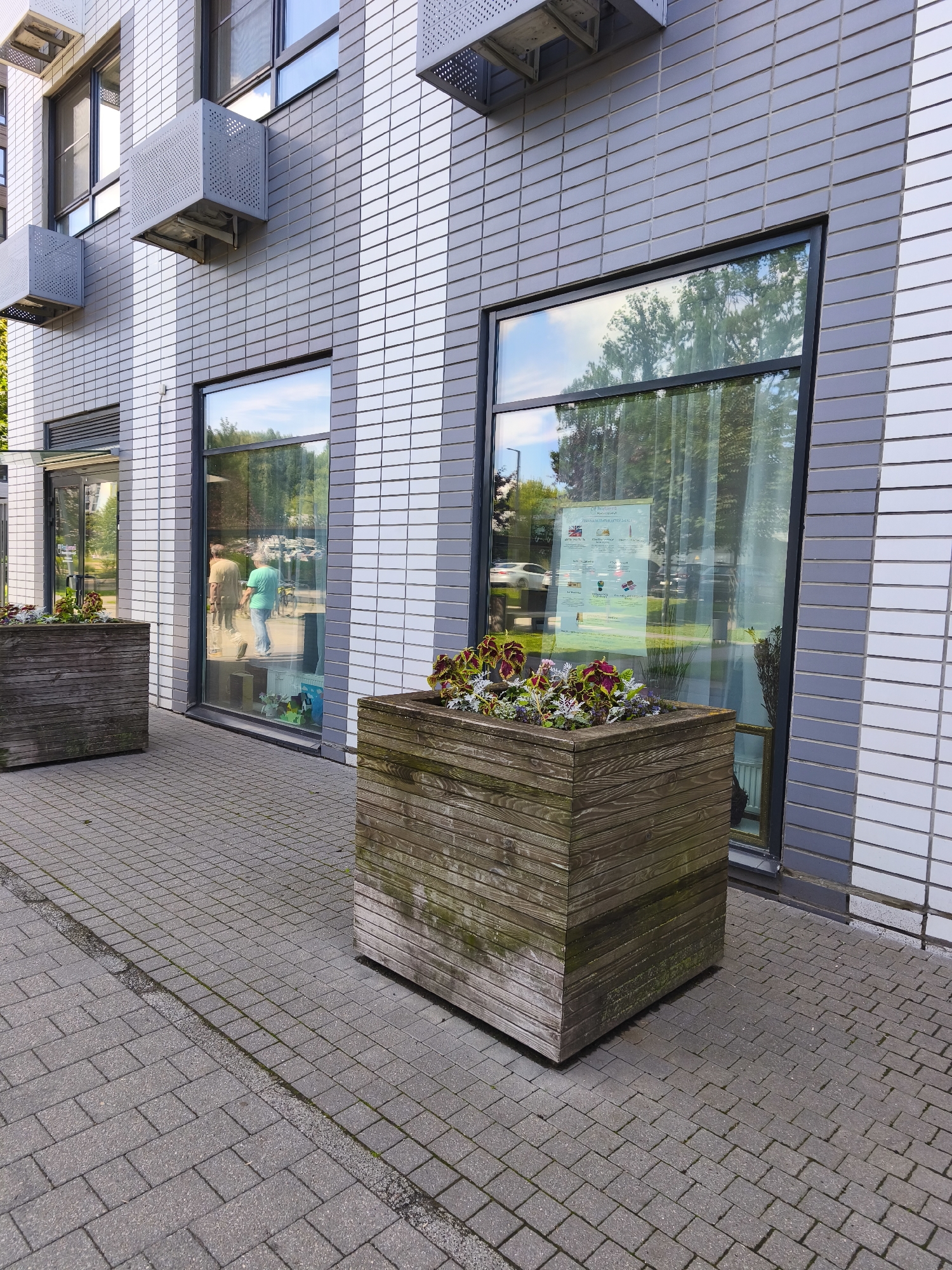 The image shows the exterior of a modern building with a tiled facade. There are large windows that reflect the surrounding trees and sky. In front of the building are two large wooden planters filled with plants. The sidewalk in front of the building is paved with grey bricks. One of the windows has a poster with various flags and text. The scene appears to be a public space, possibly in front of a commercial building or office. There are no visible indicators of this location being in Moscow, Russia.