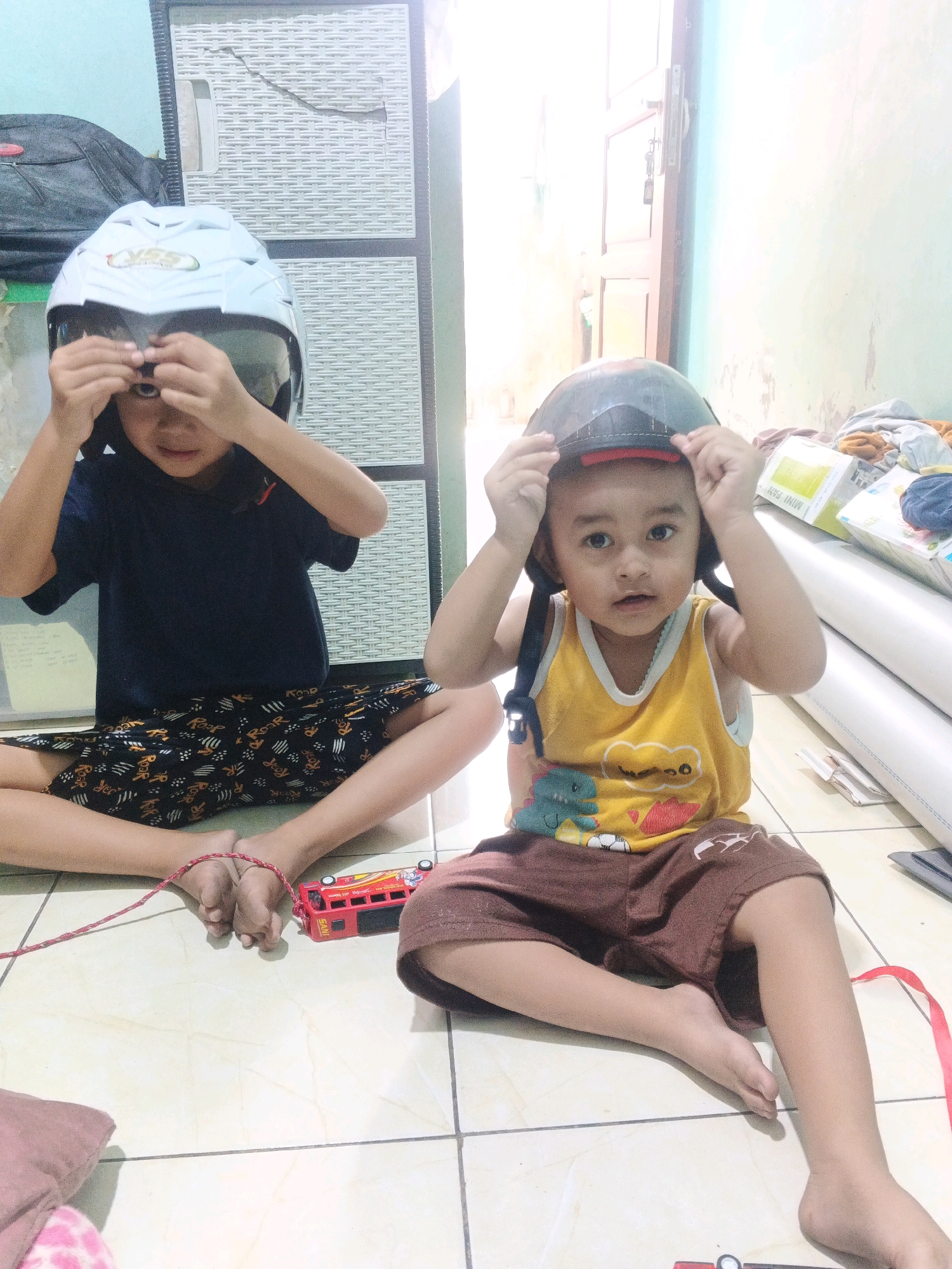 Two young children are sitting on a tiled floor indoors, playing with helmets. The child on the left, appearing to be older, wears a dark blue t-shirt and patterned shorts, with a white motorcycle helmet covering their head. They are adjusting the helmet with their hands. The child on the right, younger, is wearing a yellow tank top with a dinosaur graphic and brown shorts. A dark helmet is placed on their head, and they are also touching it. A red toy bus is visible near the feet of the child on the left, whose ankles are tied together with a red string. The background shows a white plastic cabinet and a doorway. The lighting suggests it is daytime. The scene is likely a residential interior in Kadu, Indonesia.